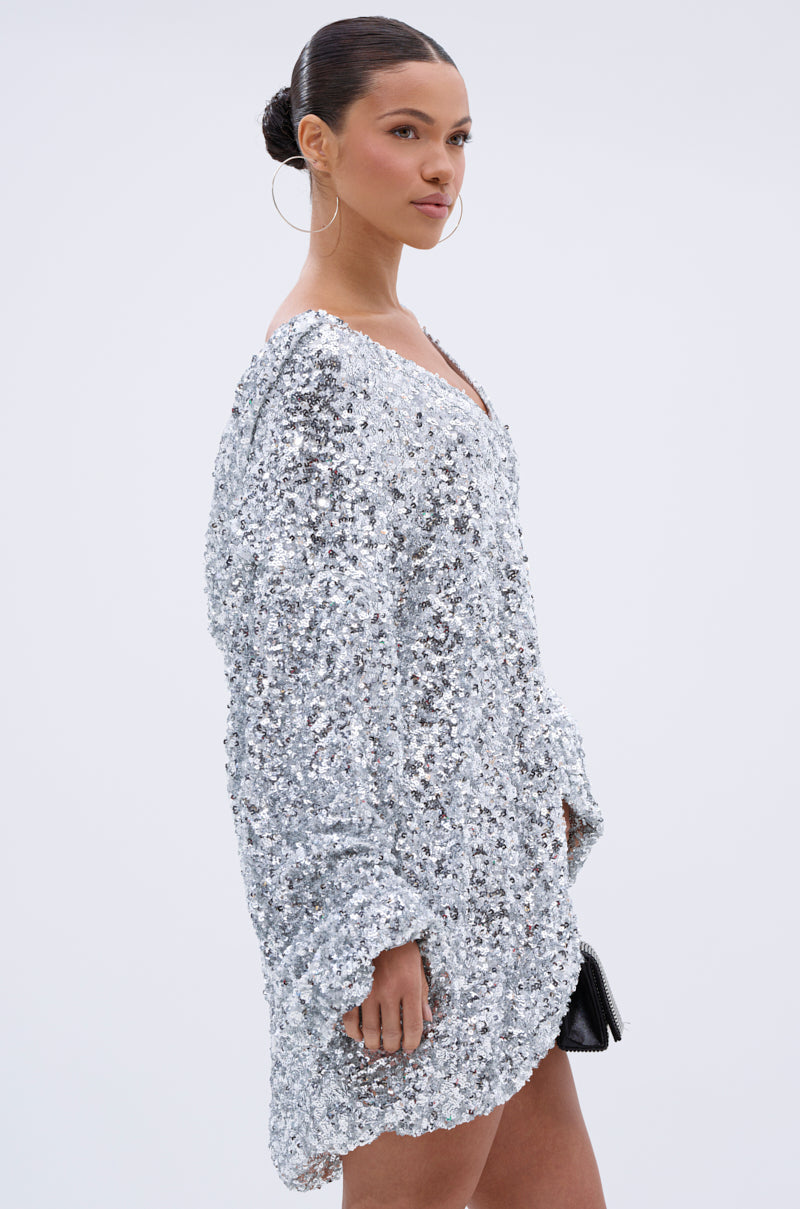 A woman with slicked-back hair and large hoop earrings models the GISELLE SEQUIN SWEATSHIRT DRESS IN SILVER, pairing it with a small black clutch against a plain light background.