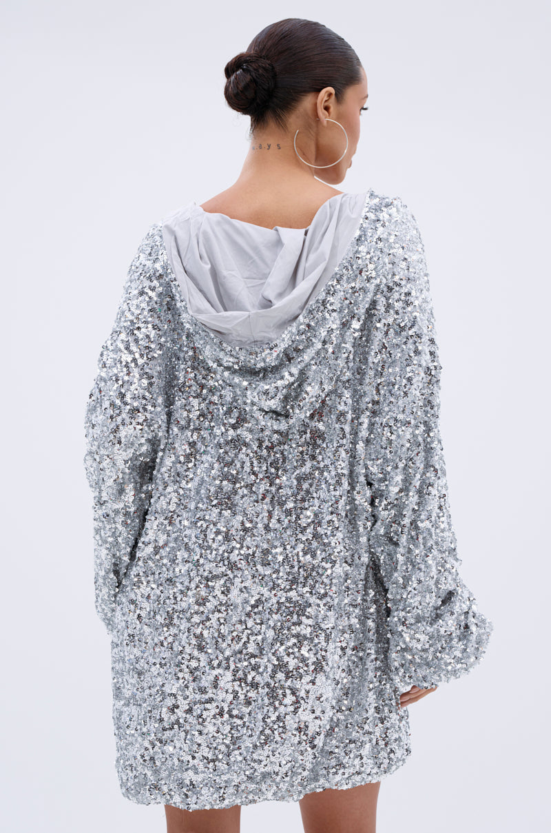 A woman with her hair in a bun and large hoop earrings models the GISELLE SEQUIN SWEATSHIRT DRESS IN SILVER, standing with her back to the camera against a plain light background.