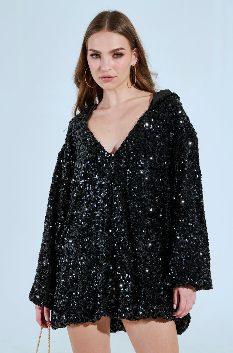 Set against a light blue backdrop, a person dazzles in the GISELLE SEQUIN SWEATSHIRT DRESS with shimmering sequin embellishments. Long brown hair flows down as they clutch a small gold strap bag. The dress boasts a deep V-neck and long sleeves, radiating elegance and allure. 1