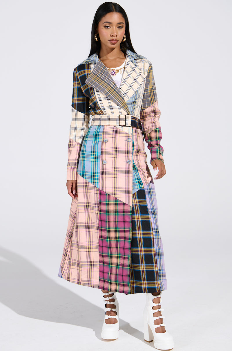 A woman poses against a light background wearing the LOGAN PATCHWORK TRENCH, a long, multicolored plaid coat with a belt, perfectly accentuating her white platform heels and long dark hair, effortlessly capturing attention.