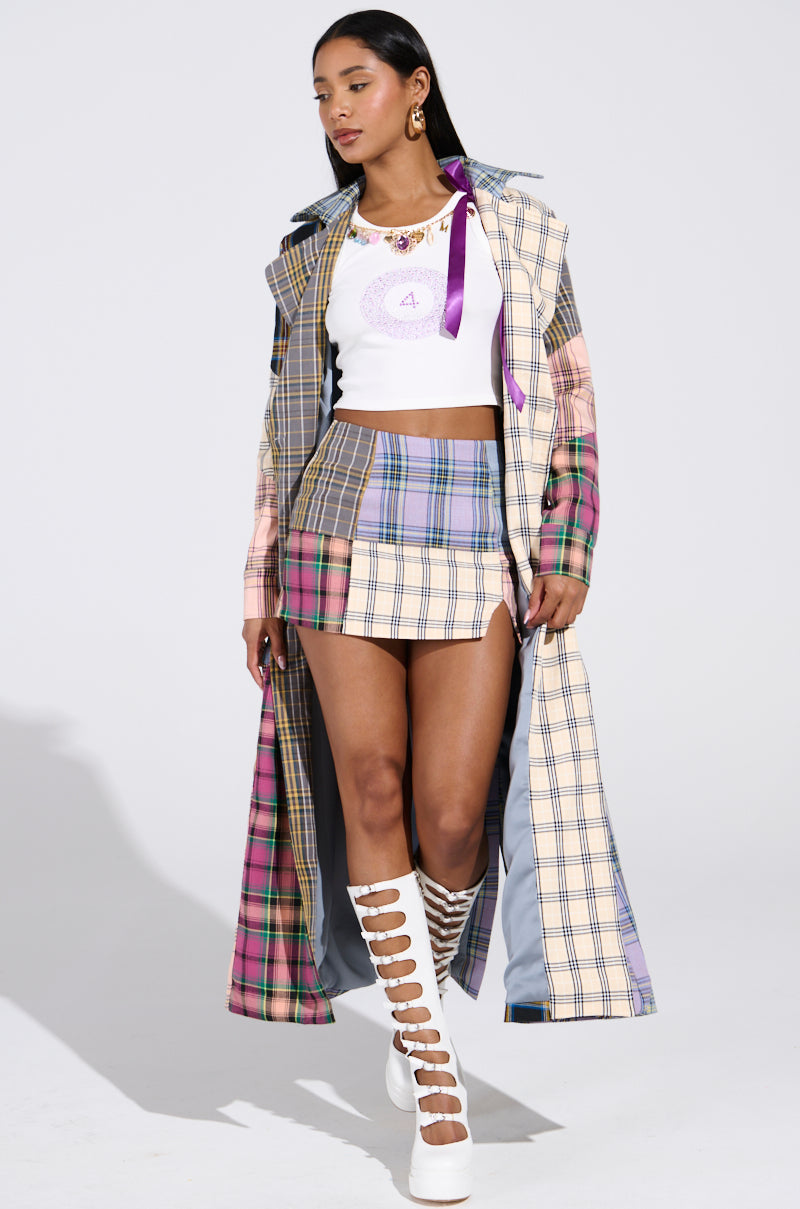 A person dons the LOGAN PATCHWORK TRENCH featuring pink, green, and blue tones over a white crop top and Logan patchwork skirt. Their long dark hair complements the ensemble, paired perfectly with white knee-high lattice-style boots.