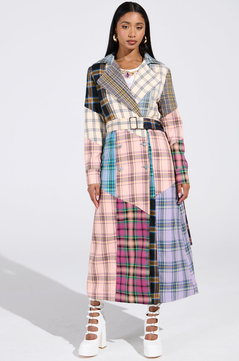 A woman wears the LOGAN PATCHWORK TRENCH, a long belted coat with vibrant plaid hues of beige, blue, pink, purple, and green. She pairs it with white knee-high platform boots against a plain background.