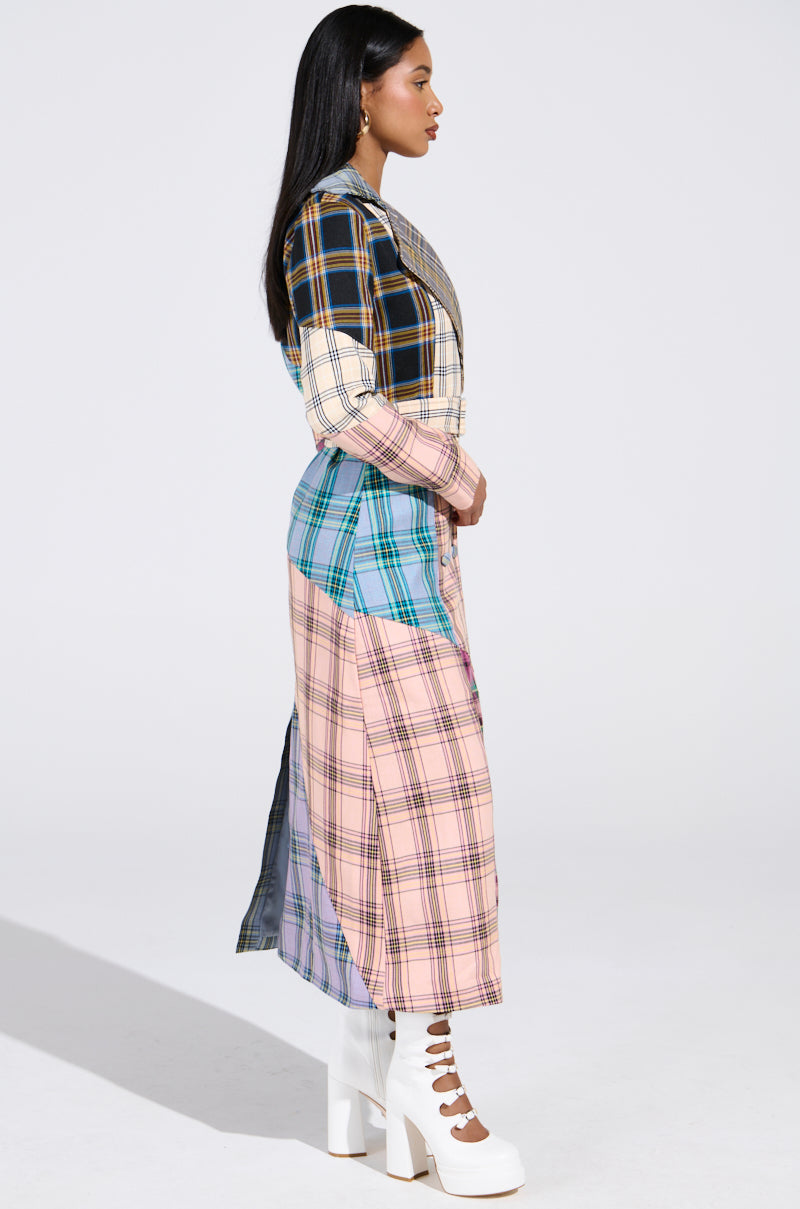 A person with long hair stands in profile wearing a vibrant LOGAN PATCHWORK TRENCH, featuring various colors including black, white, pink, and blue. They pair it elegantly with white heeled boots against a plain white background.