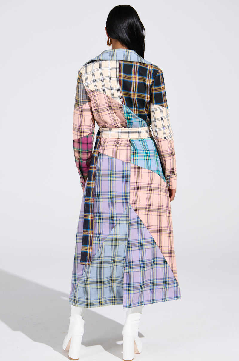 A person is seen from the back wearing the LOGAN PATCHWORK TRENCH, a long coat combining pastel and dark plaid patterns with a matching belt, paired stylishly with white heeled boots.