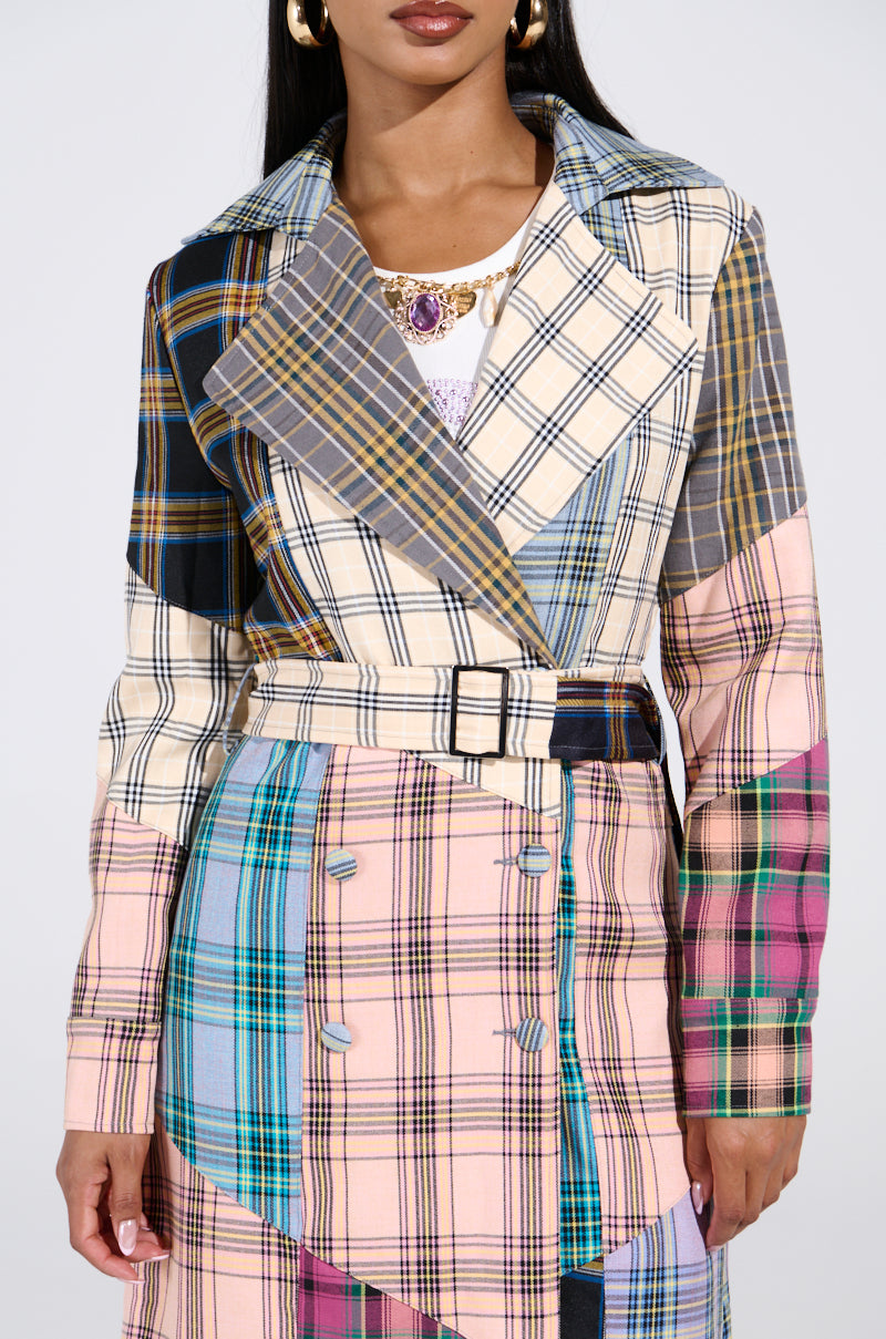 Someone wearing the vibrant LOGAN PATCHWORK TRENCH with a belted waist showcases a blend of blue, pink, yellow, and green check patterns. They have long dark hair, paired with gold hoop earrings and a jeweled necklace.