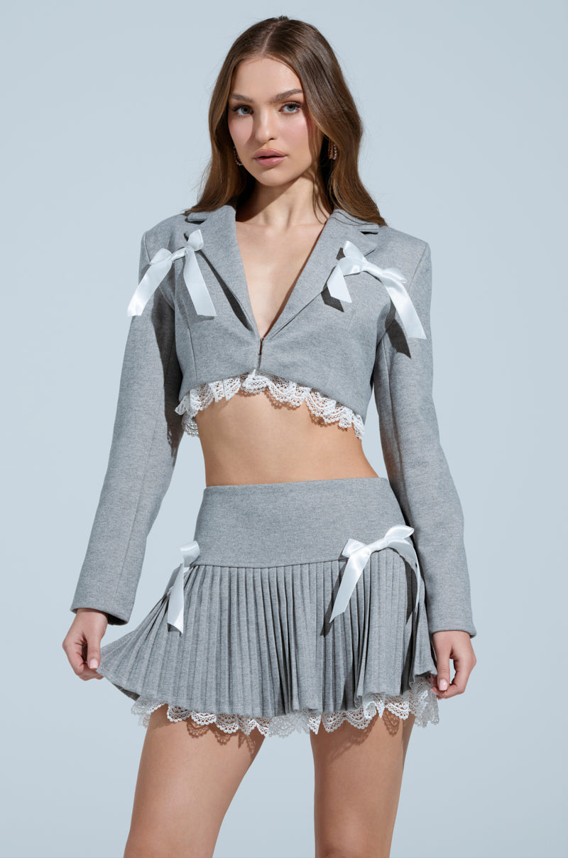 A woman with long hair poses against a light blue background in a gray two-piece outfit featuring white lace trim and satin bows. The cropped top, similar to the MARLEY LACE TRIM BLAZER with a notch-lapel collar, pairs with a pleated mini skirt, complementing her neutral expression.