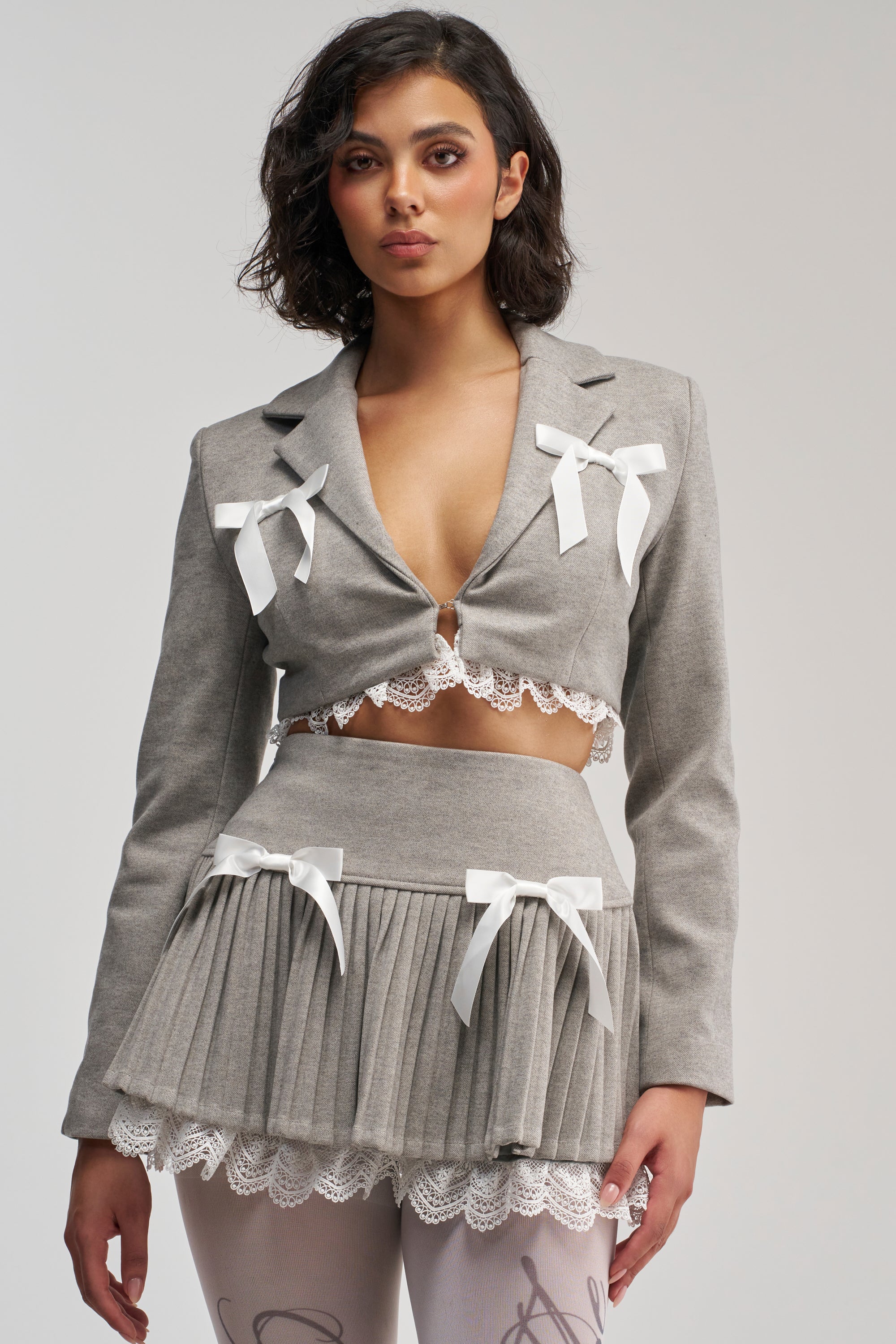 A woman poses in the MARLEY LACE TRIM BLAZER, a gray cropped piece featuring pleated details, white lace trim, and delicate white bow accents, embodying a coquette jacket-inspired look as she stands against a plain light background.