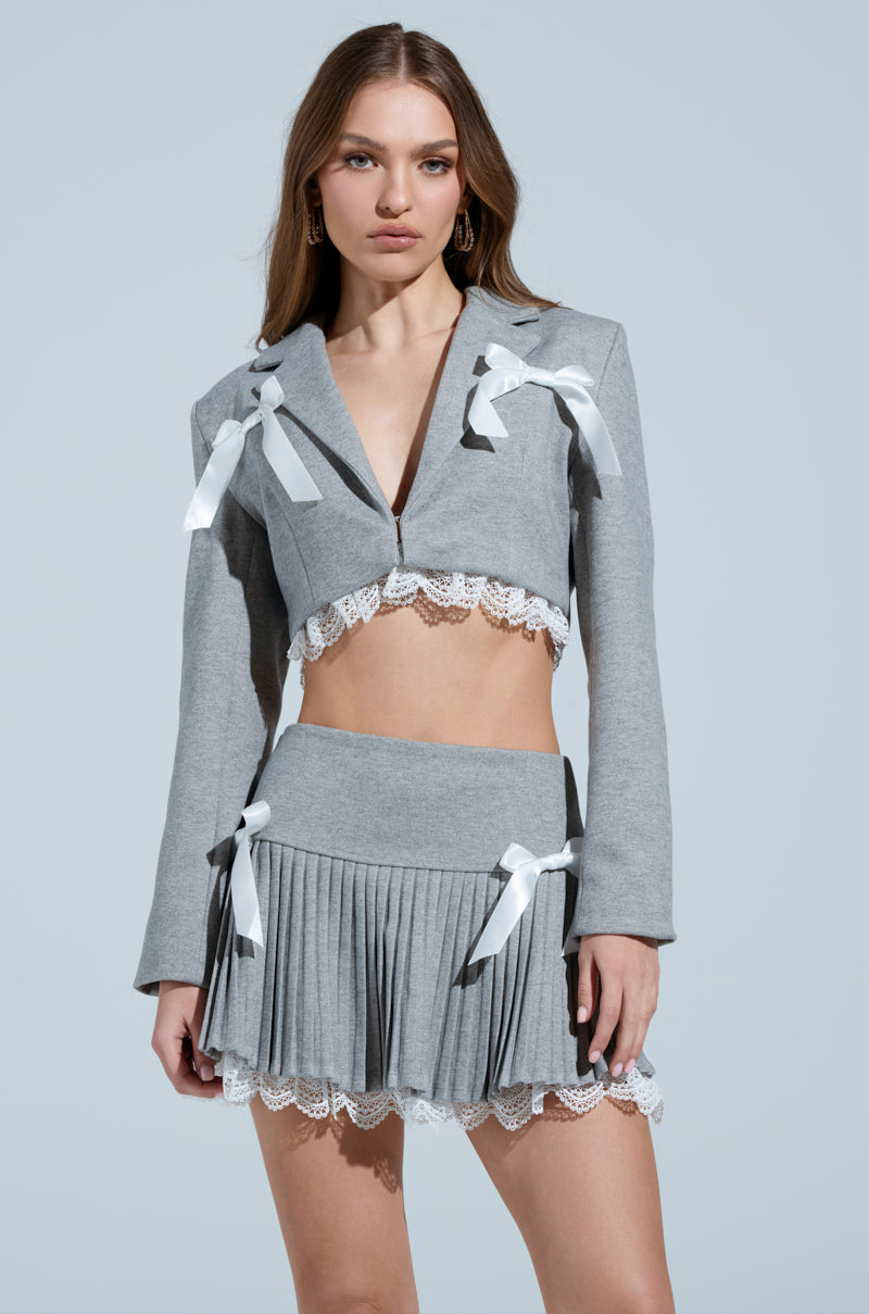 A woman with long hair sports a grey cropped Marley Lace Trim Blazer featuring white ribbon and decorative buttons, paired with a grey pleated skirt. She stands against a light blue background.