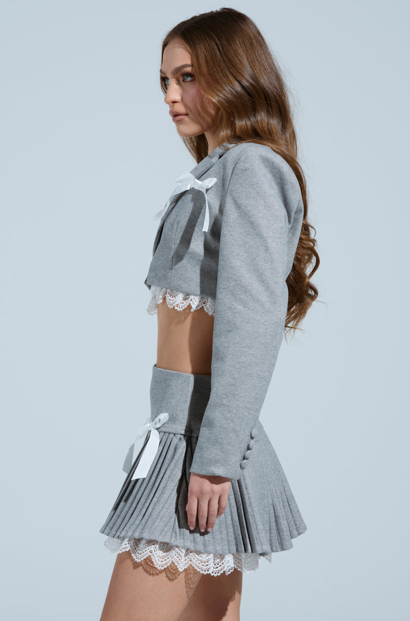 A person with long brown hair models the MARLEY LACE TRIM BLAZER—a gray cropped silhouette with a notch-lapel collar—and a pleated mini skirt with white lace trim and bows, standing sideways against a pale blue background.