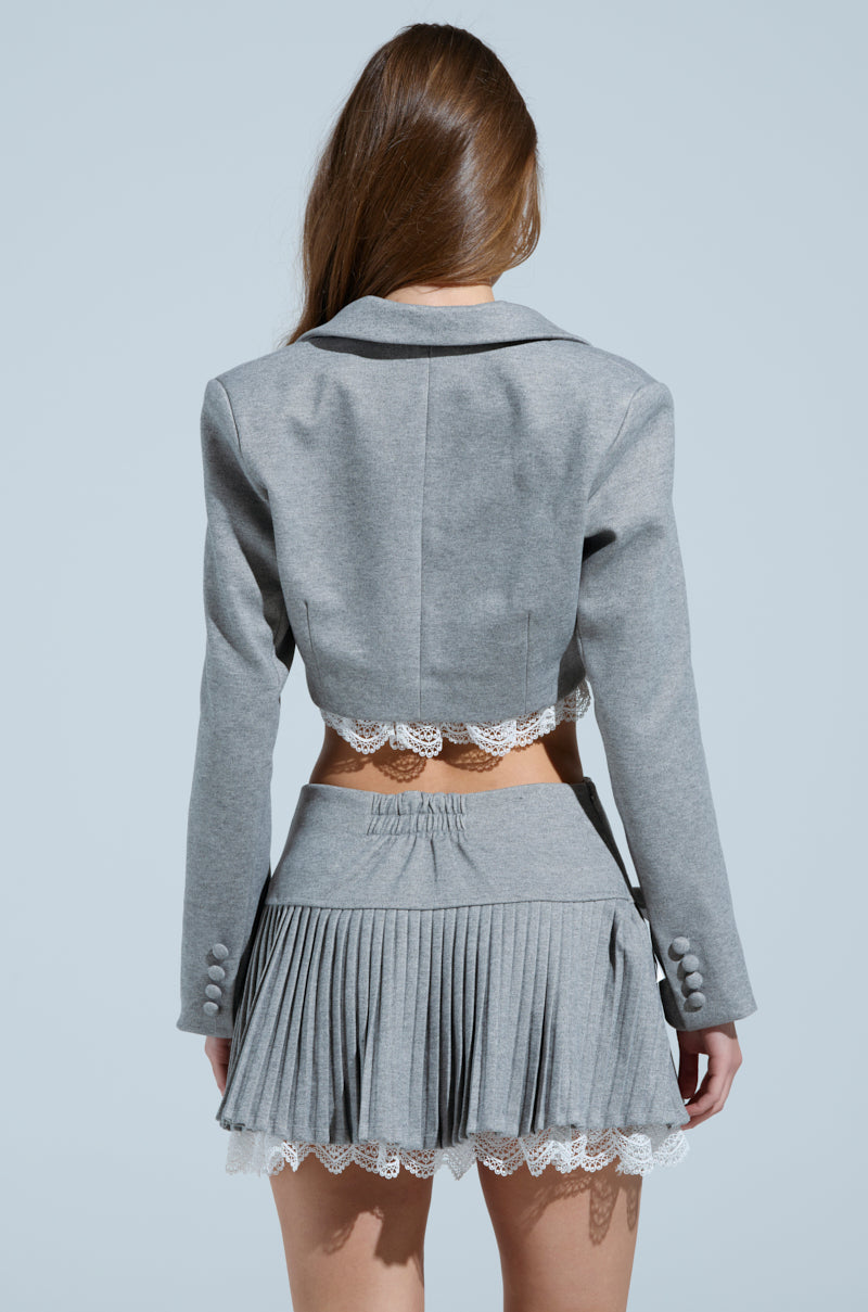 A woman faces away, highlighting the MARLEY LACE TRIM BLAZER with a grey cropped silhouette and notch-lapel collar, paired with a pleated skirt featuring lace accents. The backdrop is a calming light blue.
