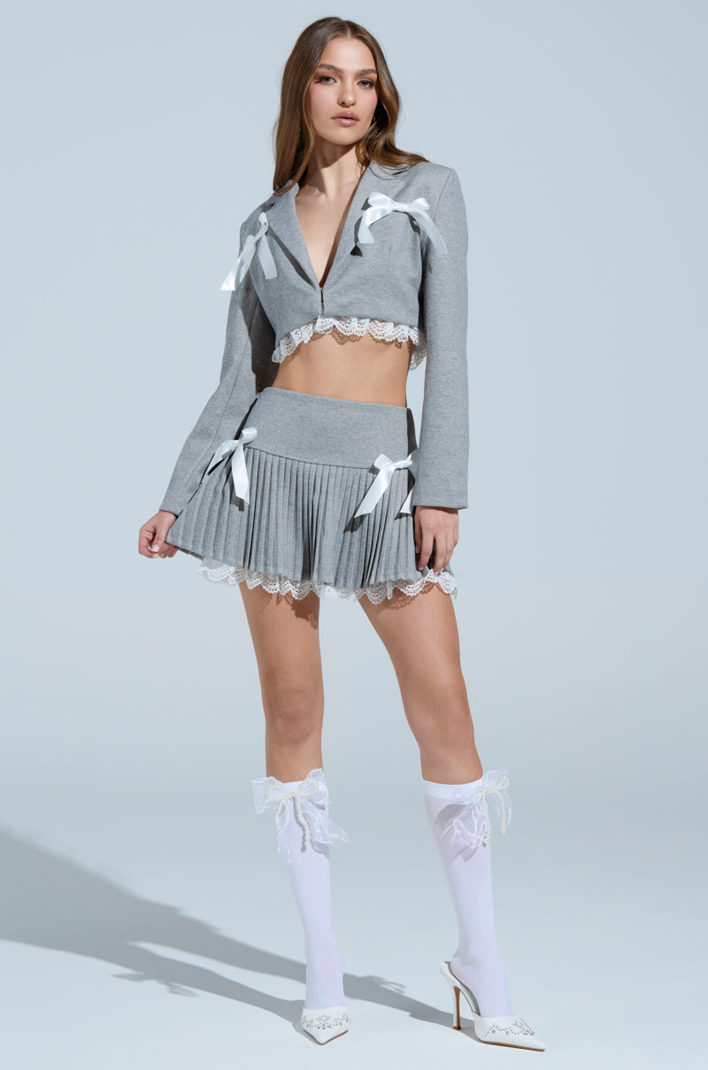 A woman poses confidently against a light blue backdrop, wearing a gray MARLEY LACE TRIM BLAZER with white bows and a notch-lapel collar. The cropped jacket complements her matching pleated skirt, knee-high socks adorned with bows, and white heels. She rests her hand on her hip.