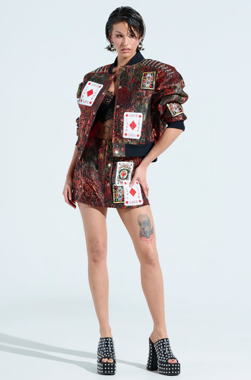 A person confidently poses in a high-rise silhouette wearing the SHOW YOUR CARDS BROCADE MINI SKIRT in red and black, adorned with playing card patches. They have short, wet-look hair, a thigh tattoo, and studded platform shoes. The backdrop is plain white.