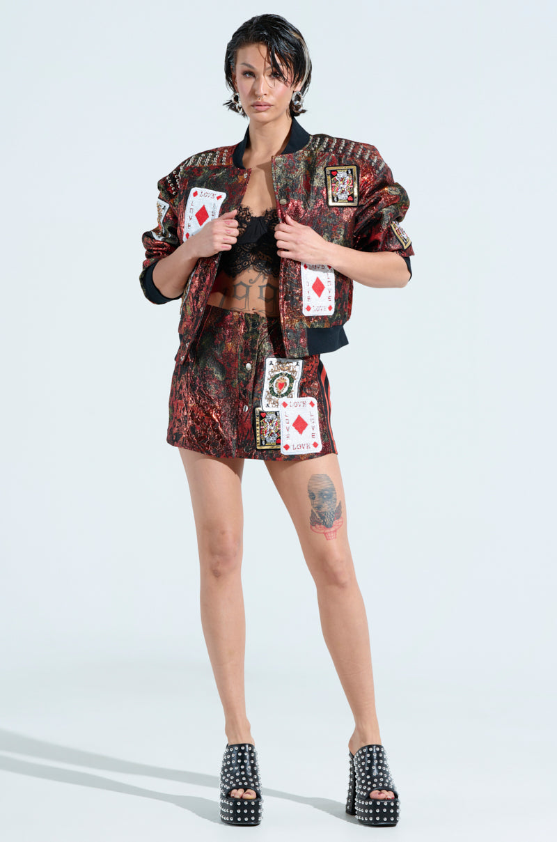 A person in a colorful outfit poses confidently, wearing a lace top, the "SHOW YOUR CARDS BROCADE MINI SKIRT," and a jacket adorned with playing card patches. Platform heels and a skull tattoo on their thigh accentuate the edgy style.