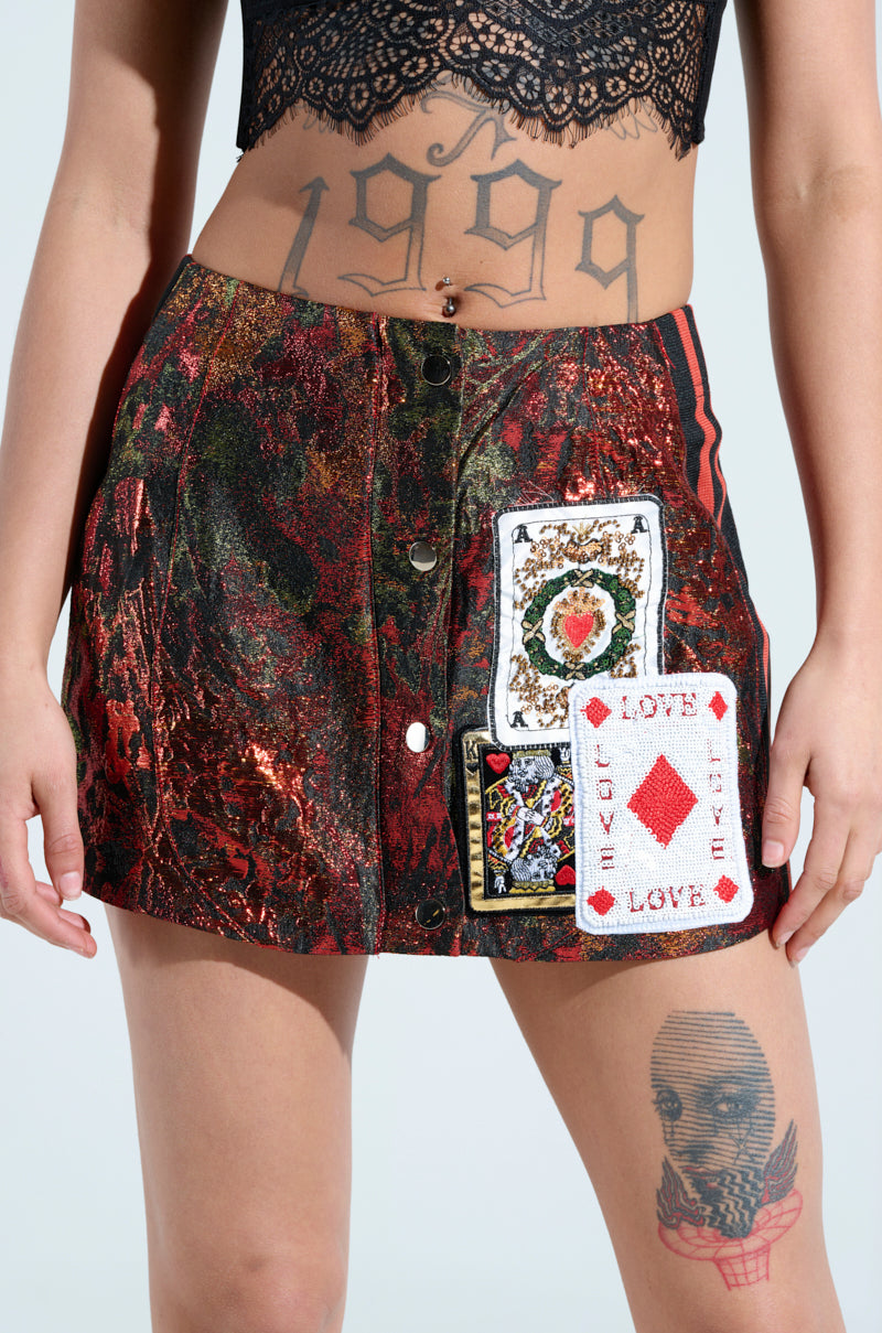 A person in a black lace top and the SHOW YOUR CARDS BROCADE MINI SKIRT with large playing card patches. The number "1999" is tattooed above the navel, accompanied by a masked face tattoo on the thigh, creating an intriguing high-rise silhouette.
