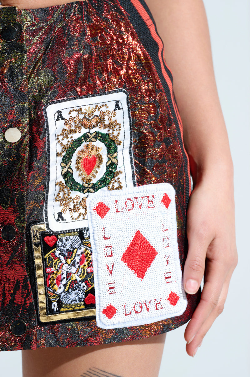 Wearing the "SHOW YOUR CARDS BROCADE MINI SKIRT," adorned with textured playing card patches, featuring an ace of spades and a diamond with "LOVE," a person reveals their tattoo while holding part of the high-rise silhouette.