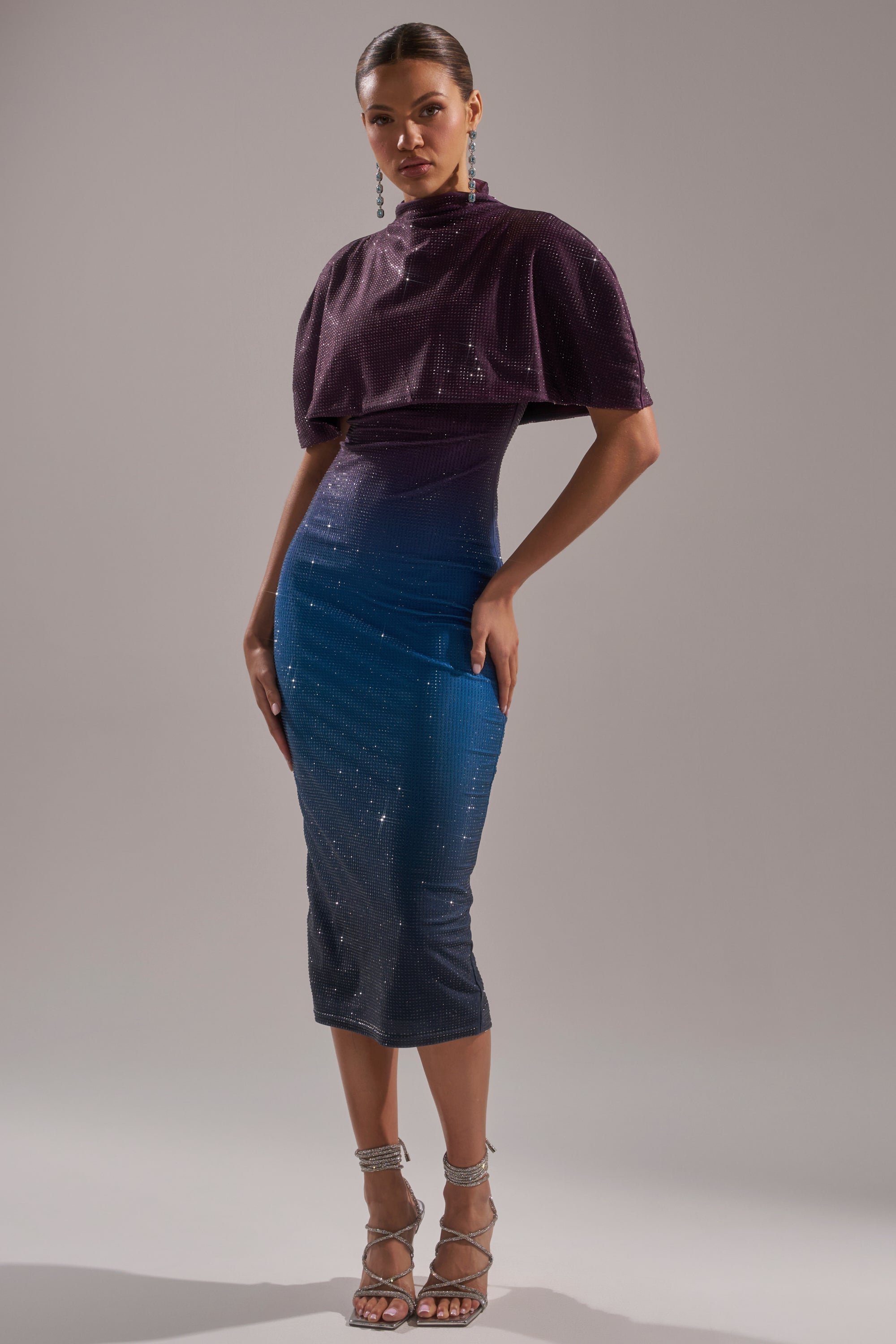 A woman models the CHIC AF RHINESTONE MIDI DRESS, a fitted sparkly design with a cape-like overlay that fades from deep purple to blue. She completes the look with strappy silver heels and long earrings against a plain background. 0
