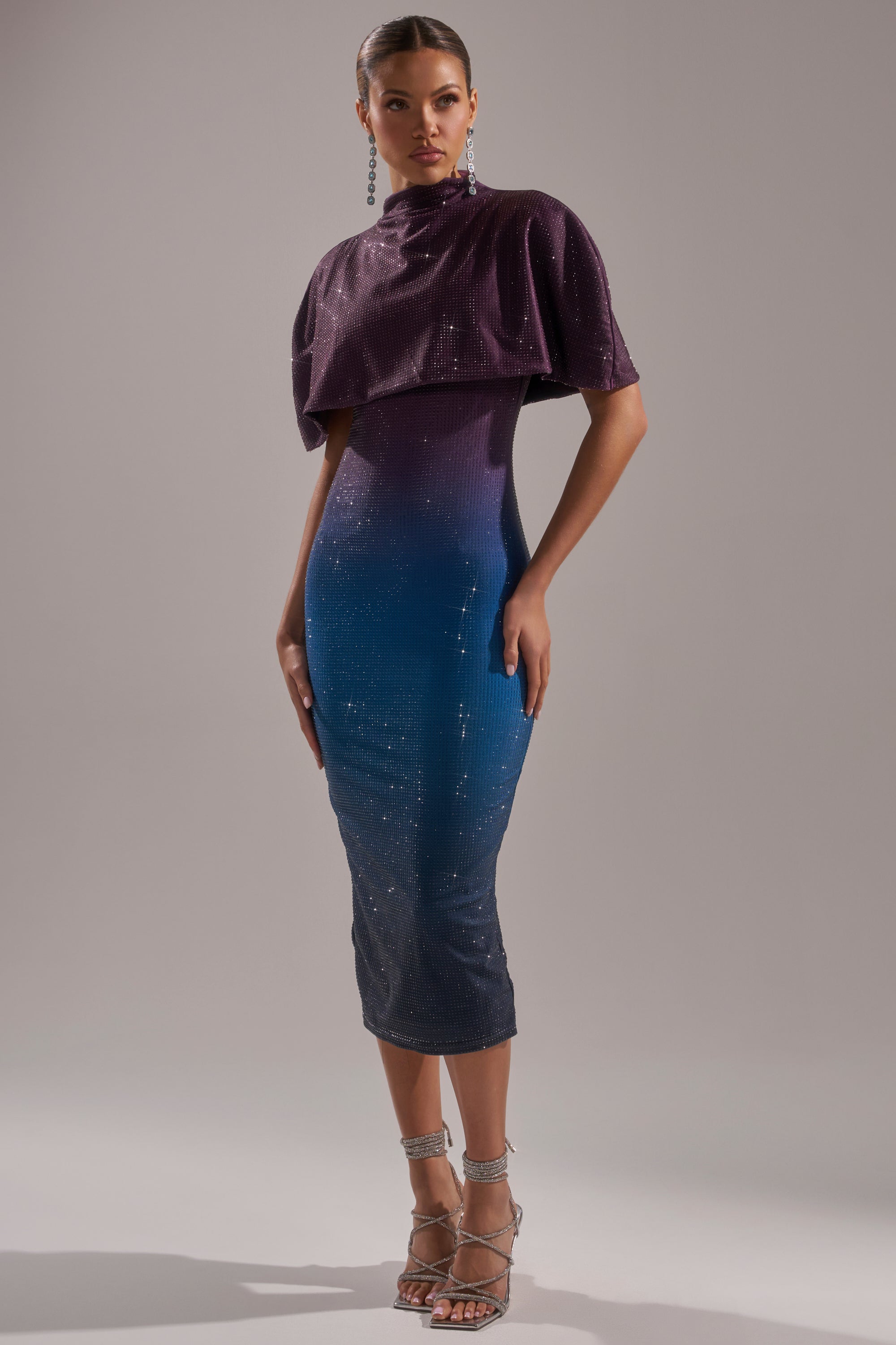 A woman models the CHIC AF RHINESTONE MIDI DRESS, a fitted, sparkly gradient piece blending deep purple to blue. She pairs it with silver strappy heels and long dangling earrings, standing against a plain background. 1