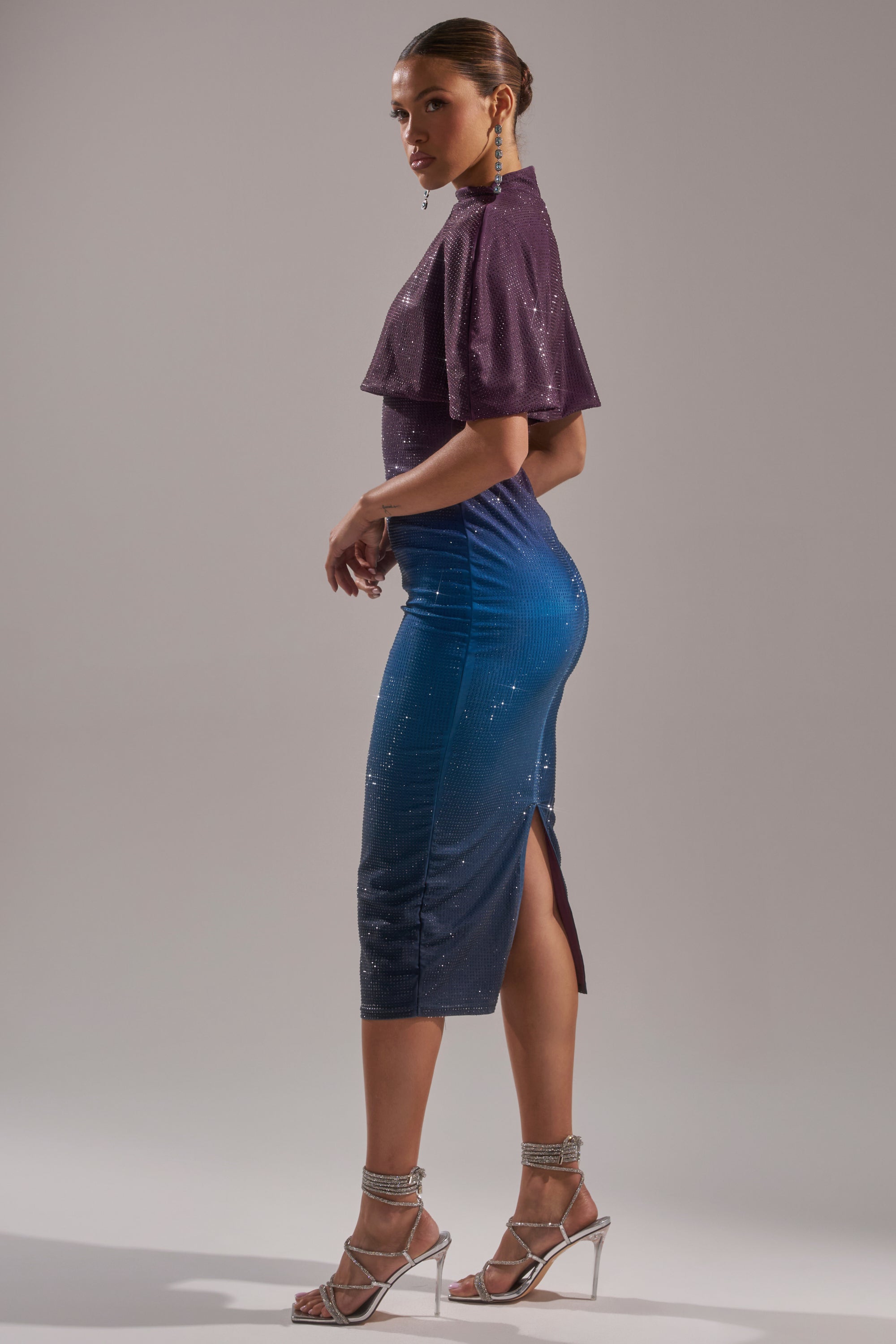 A woman poses in the CHIC AF RHINESTONE MIDI DRESS, a fitted, sparkling dress with a purple top, blue skirt, and side slit. She pairs it with silver high-heeled sandals, large dangling earrings, and wears her hair in a sleek bun. 2