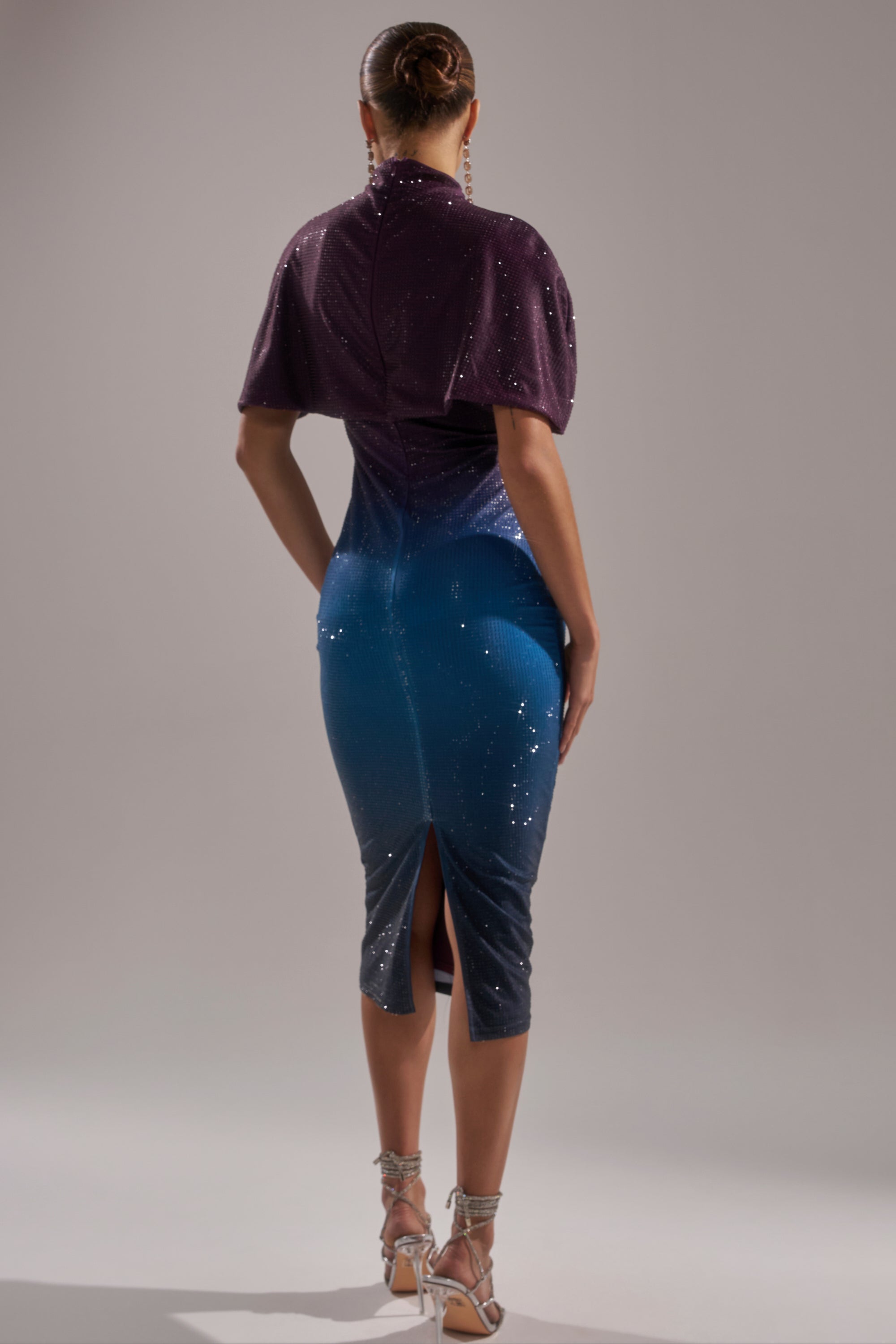 A person stands facing away, wearing the CHIC AF RHINESTONE MIDI DRESS—a sparkling, form-fitting gown that fades from deep purple to blue with a back slit. Their hair is in a bun and they wear high-heeled sandals. 3