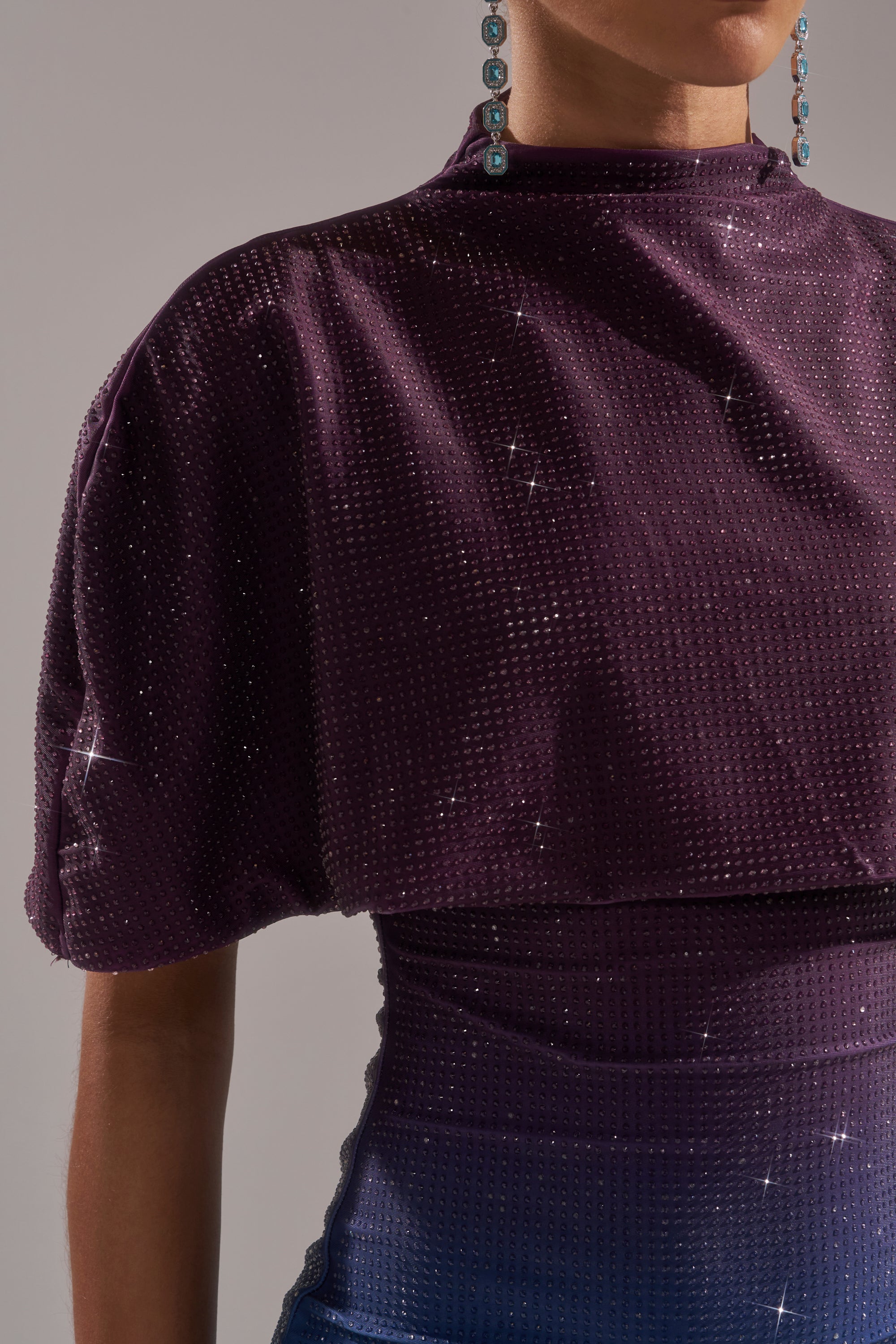 A person wearing the CHIC AF RHINESTONE MIDI DRESS in textured deep purple with shimmer; only the upper torso and right arm are shown, turquoise earrings partially visible. Background is neutral gray. 4