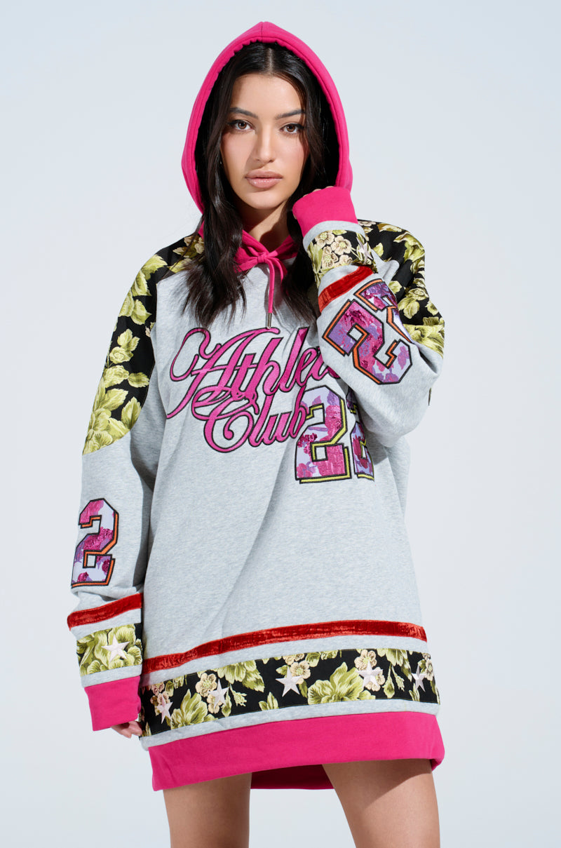 A woman wears the ATHLETIC BROCADE SWEATSHIRT DRESS: an oversized gray hoodie dress with pink and gold floral patterns, "Athletic Club 23" text on the front, and pink hood and trim. She poses against a simple, light background.