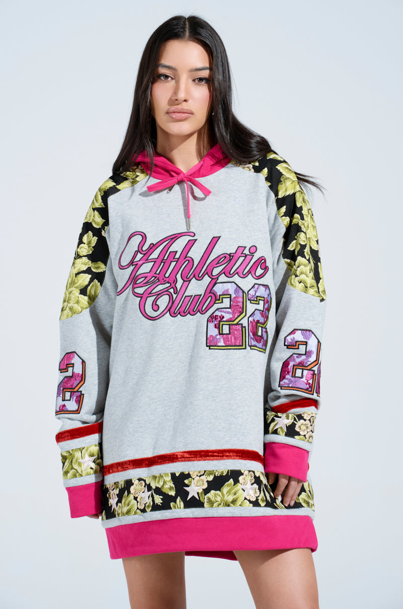 A woman in the ATHLETIC BROCADE SWEATSHIRT DRESS features a trendy oversized silhouette with floral sleeve patterns and the number "22" on each arm. "Athletic Club" is in pink and red. Her long dark hair cascades against a plain light background.