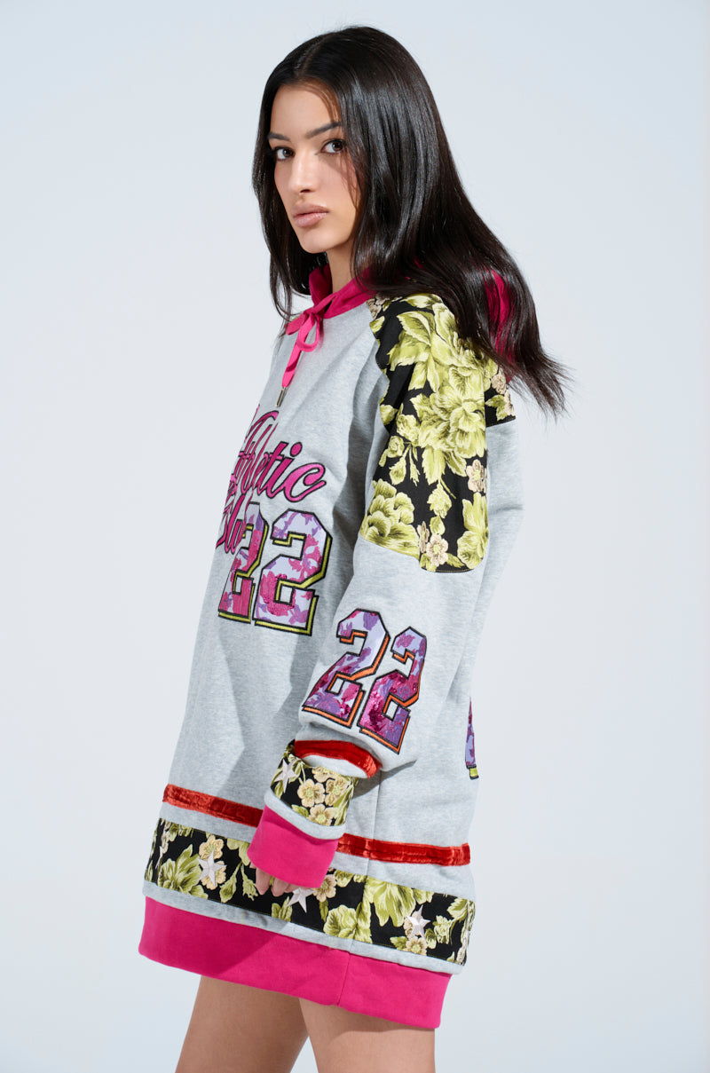 A woman poses in an ATHLETIC BROCADE SWEATSHIRT DRESS, a long, oversized hoodie featuring "22" and "Barbie." It has a gray base, pink cuffs, and yellow floral brocade on the sleeves. She stands against a simple light background.
