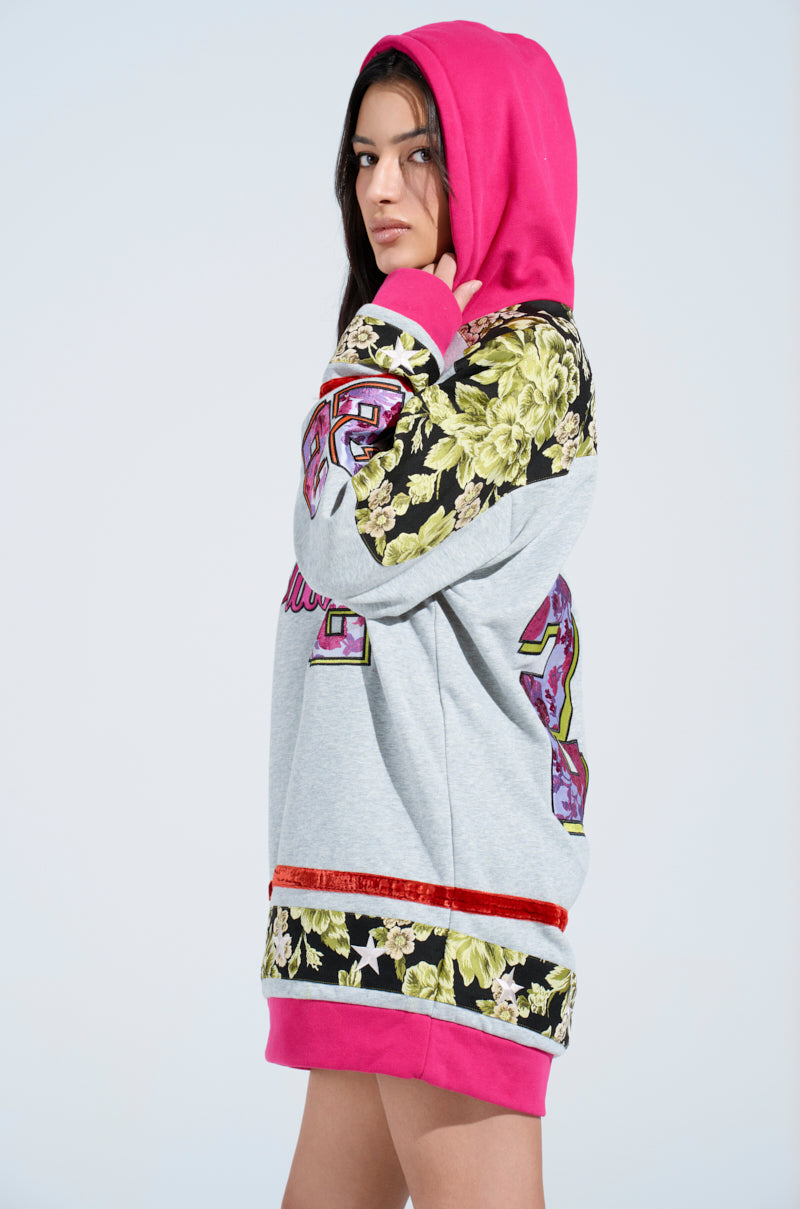 A person wearing an oversized ATHLETIC BROCADE SWEATSHIRT DRESS with a pink hood and floral brocade patterns stands against a light background. The dress features numbers and decorative designs, as the individual gazes sideways, hands resting by their sides.