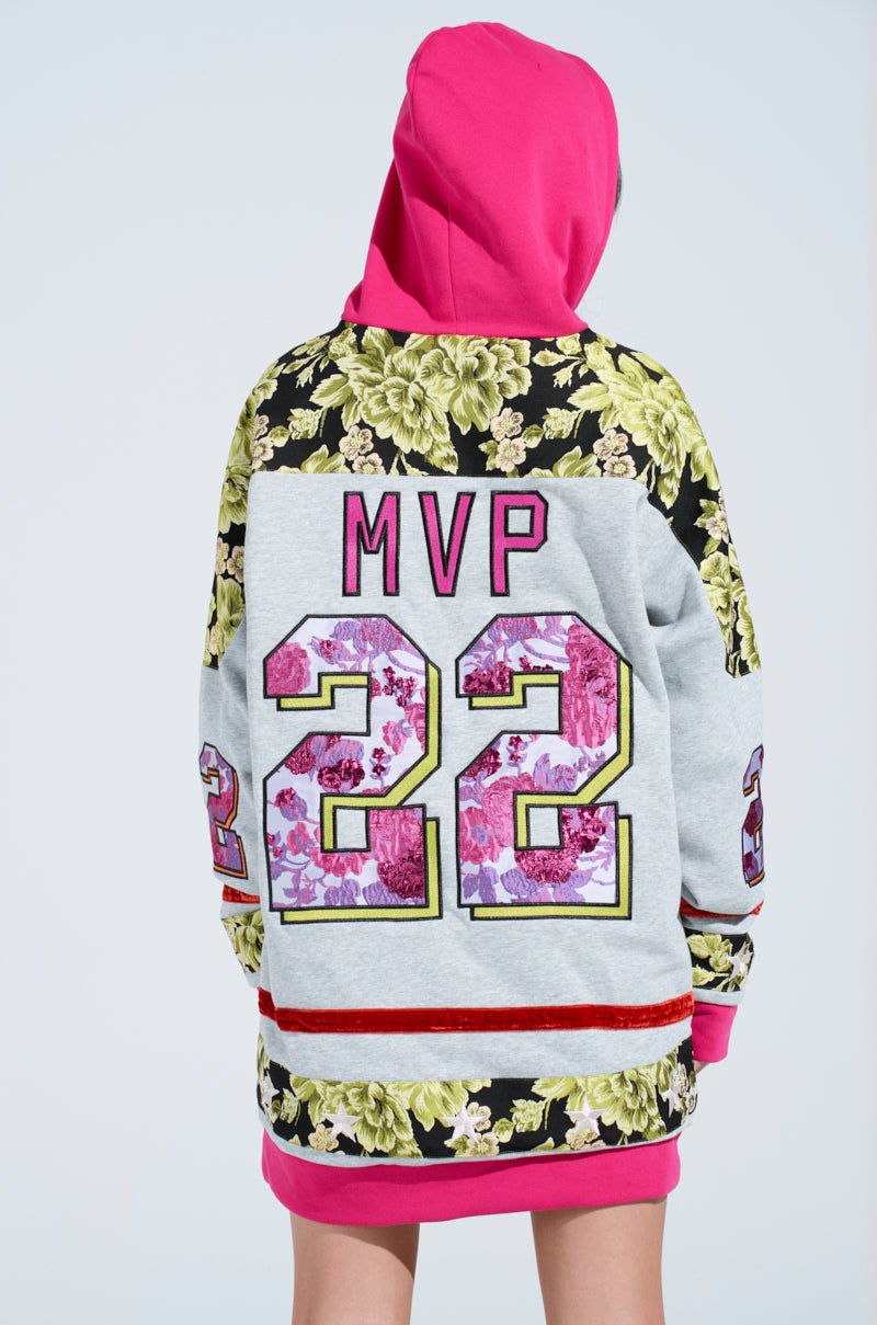 A person wears the ATHLETIC BROCADE SWEATSHIRT DRESS featuring an oversized hood with a pink "MVP" and "22" on the back. It is primarily gray with black and green floral brocade patterns, red trim, and a pink hood, as they face away.