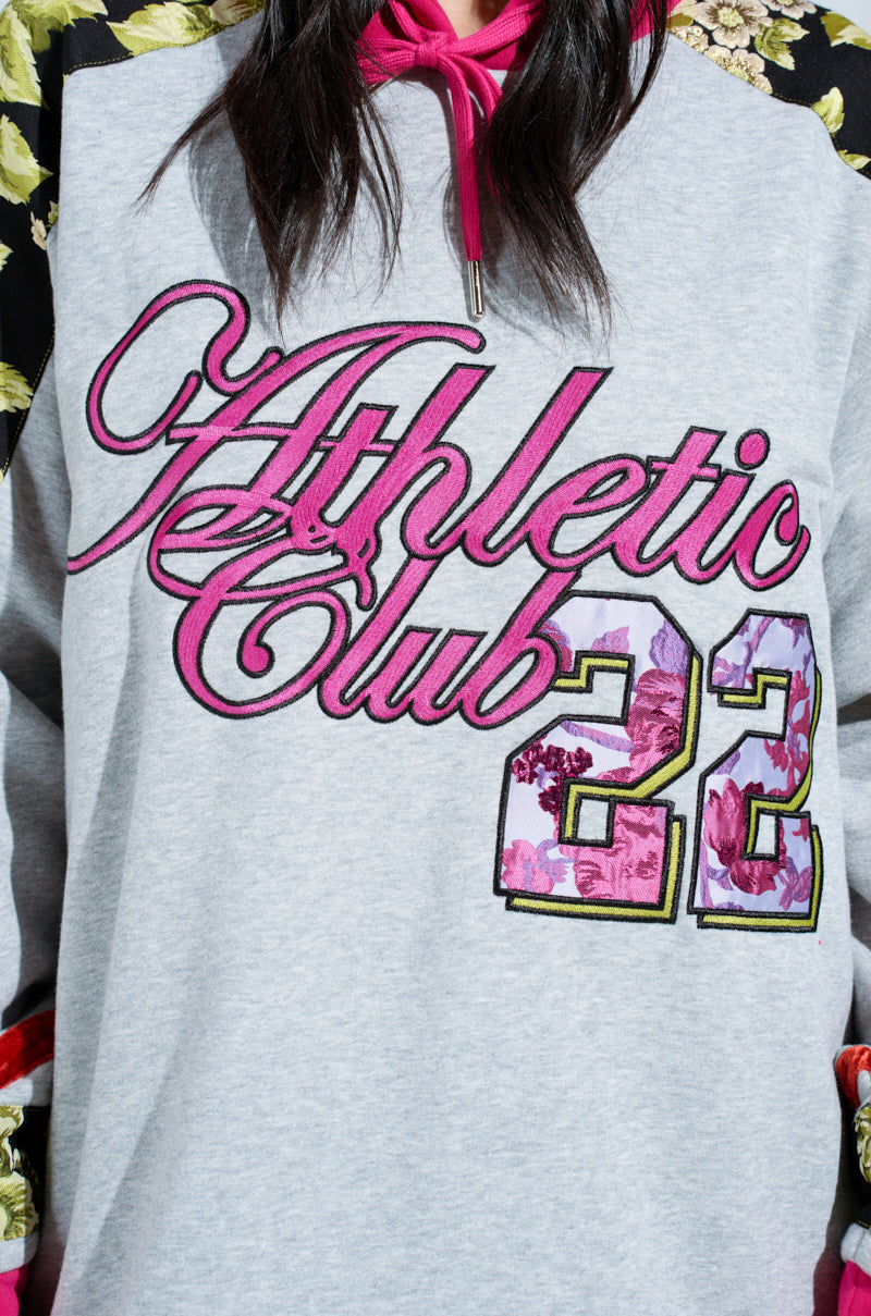 A person wears an oversized ATHLETIC BROCADE SWEATSHIRT DRESS in gray, featuring "Athletic Club 22" with pink floral accents. The sweatshirt dress has sleeves with a floral brocade design and chic pink strings on the hood.