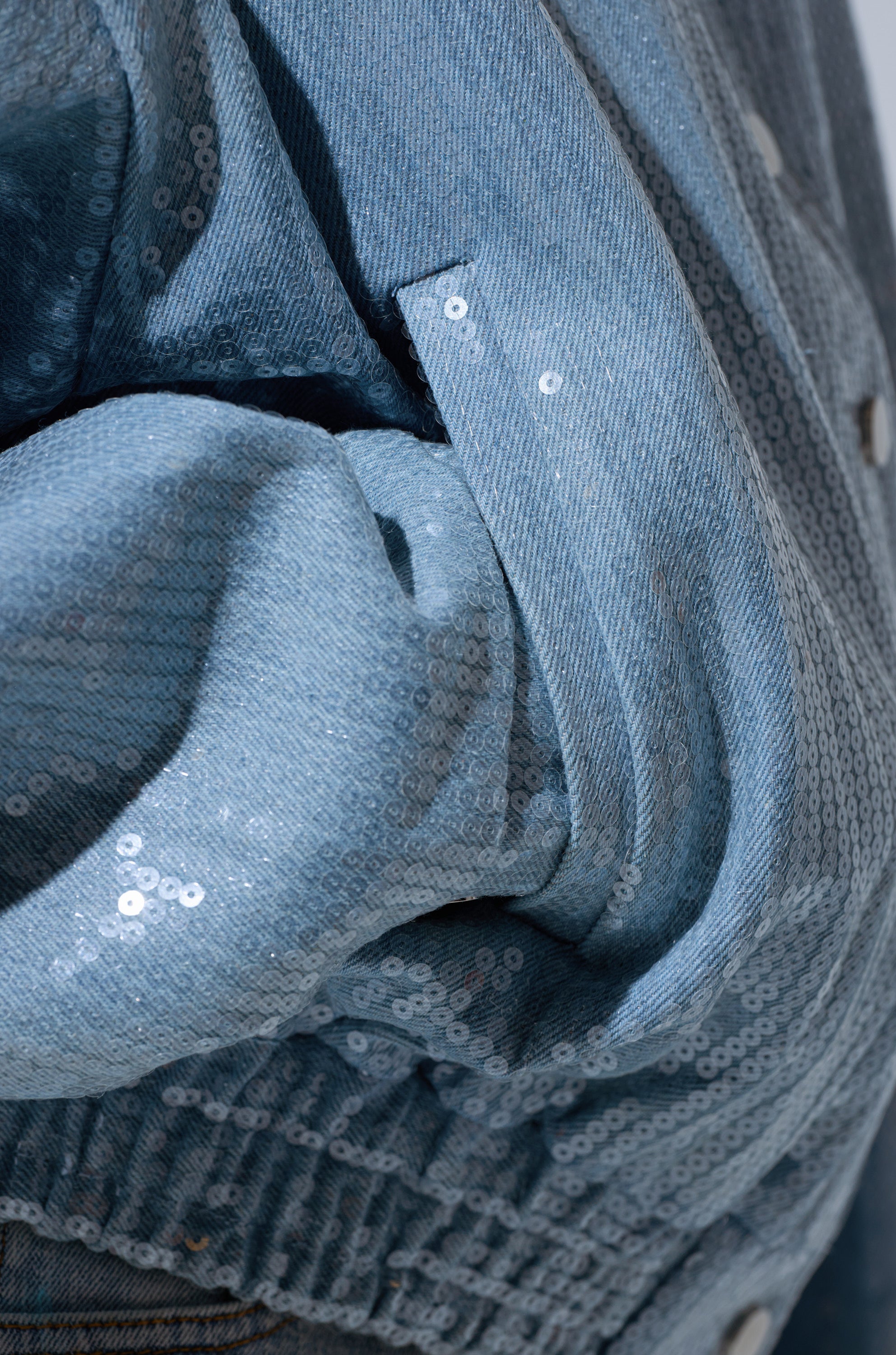 Close-up of the SEQUIN OVERLAY DENIM BOMBER in light blue denim, featuring a pocket, elastic waistband, and sparkling small sequins for a stylish, oversized look.