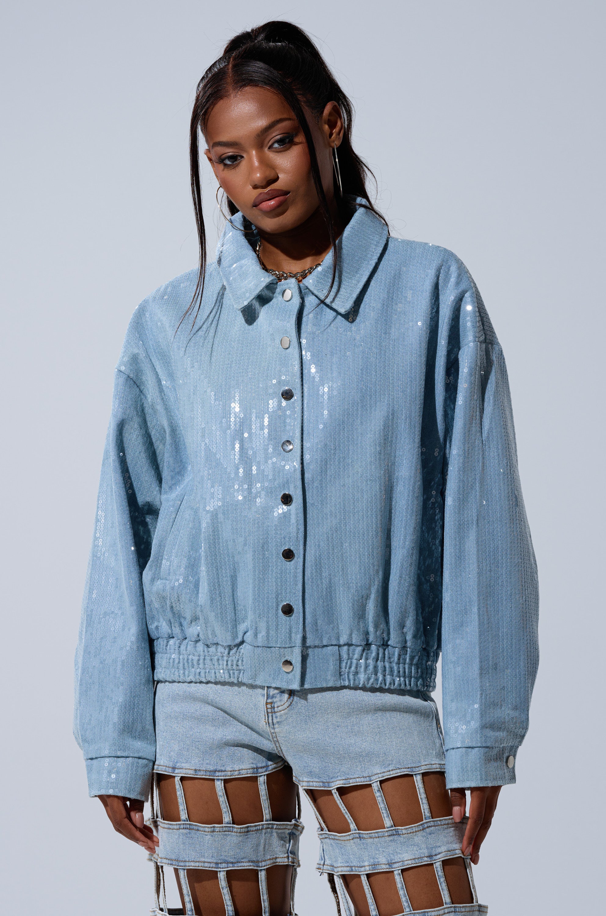 A woman models the SEQUIN OVERLAY DENIM BOMBER, a light blue sequin jacket, paired with light-wash denim shorts featuring multiple cut-outs, standing against a plain light gray background.