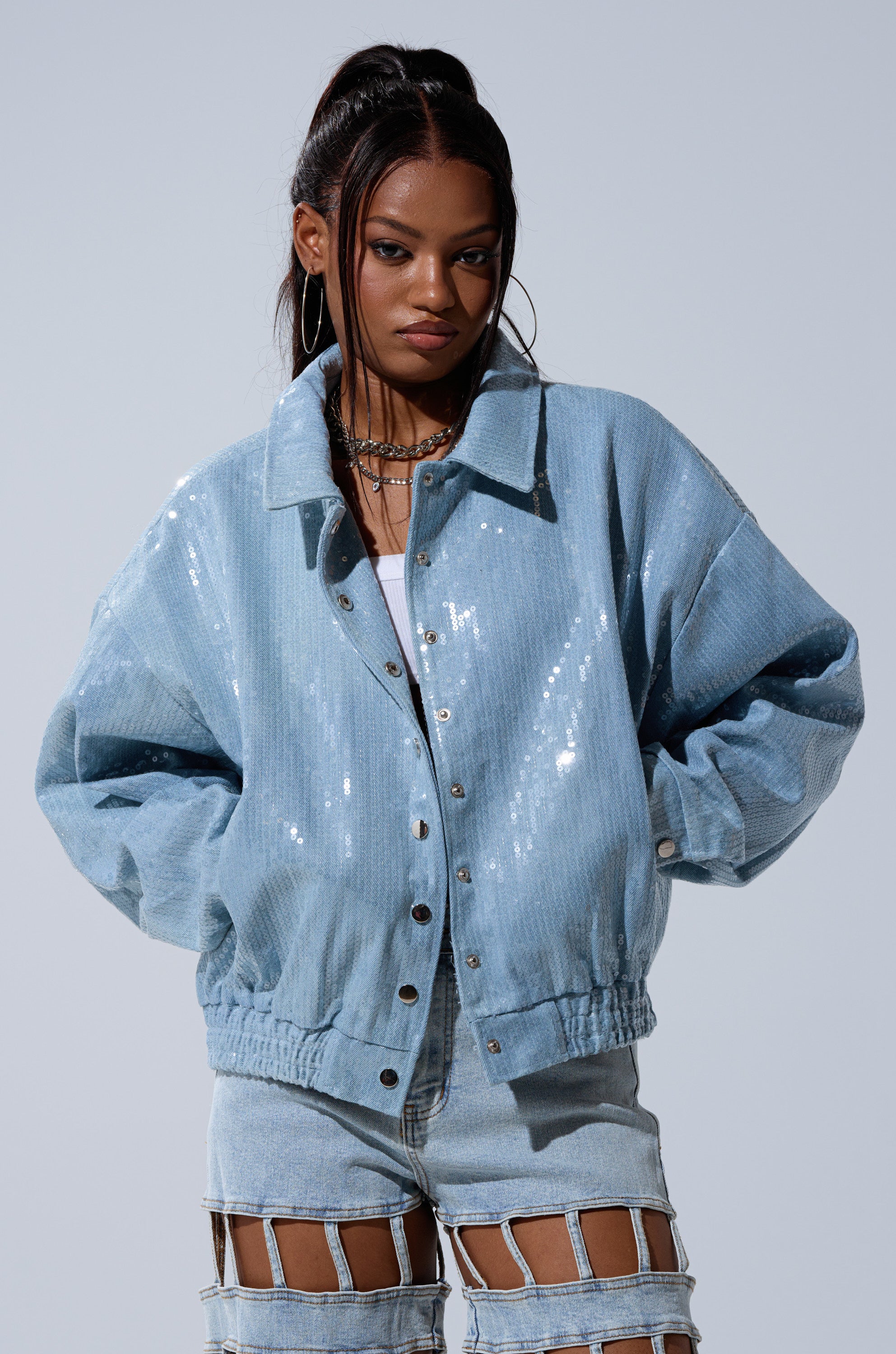 A woman stands confidently against a plain light gray background, wearing the SEQUIN OVERLAY DENIM BOMBER paired with light denim shorts featuring cutout details.