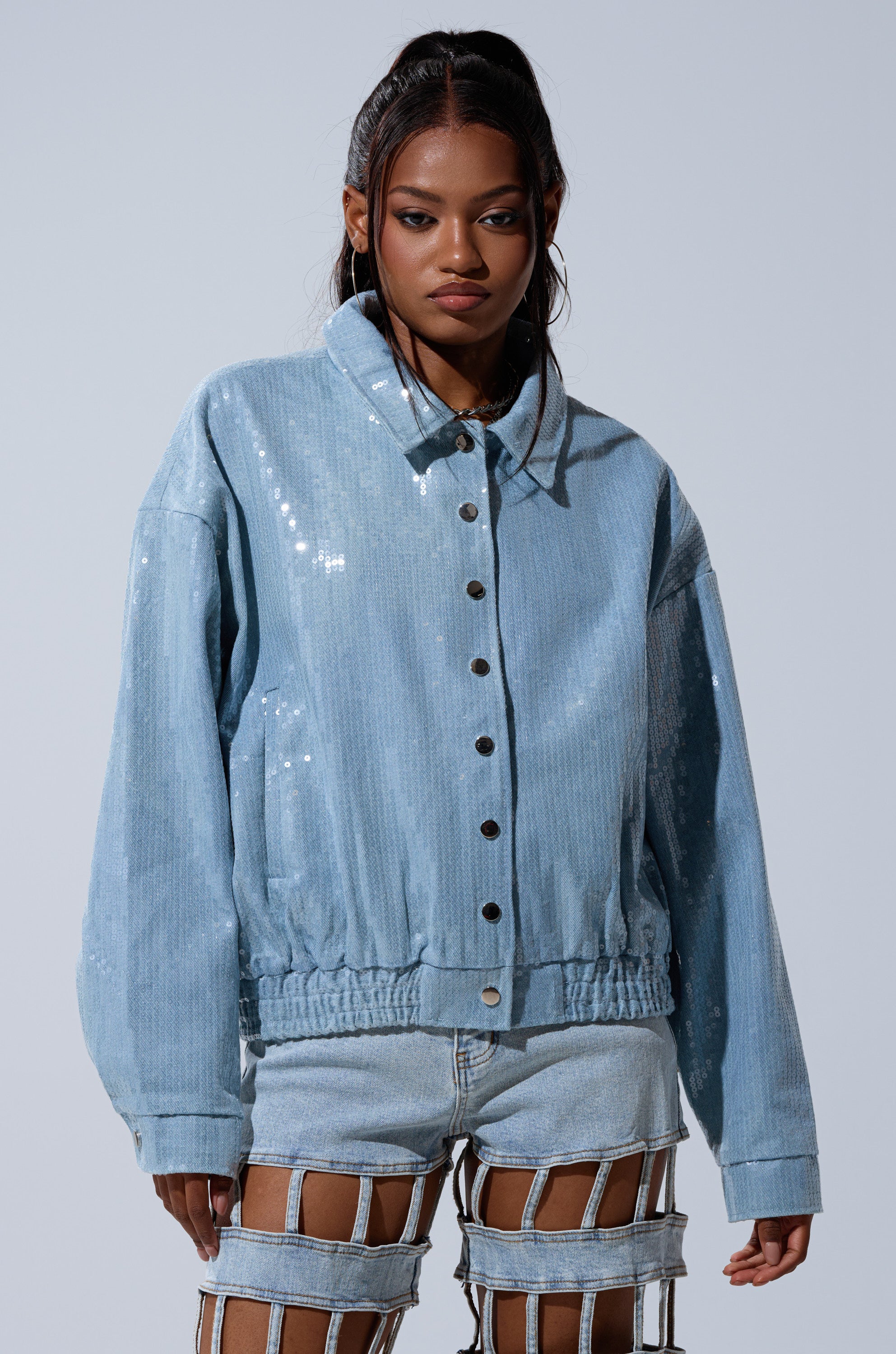 A woman wears the SEQUIN OVERLAY DENIM BOMBER, a light blue sequin jacket with silver buttons, paired with matching cutout shorts and thin straps, standing against a plain light gray background.