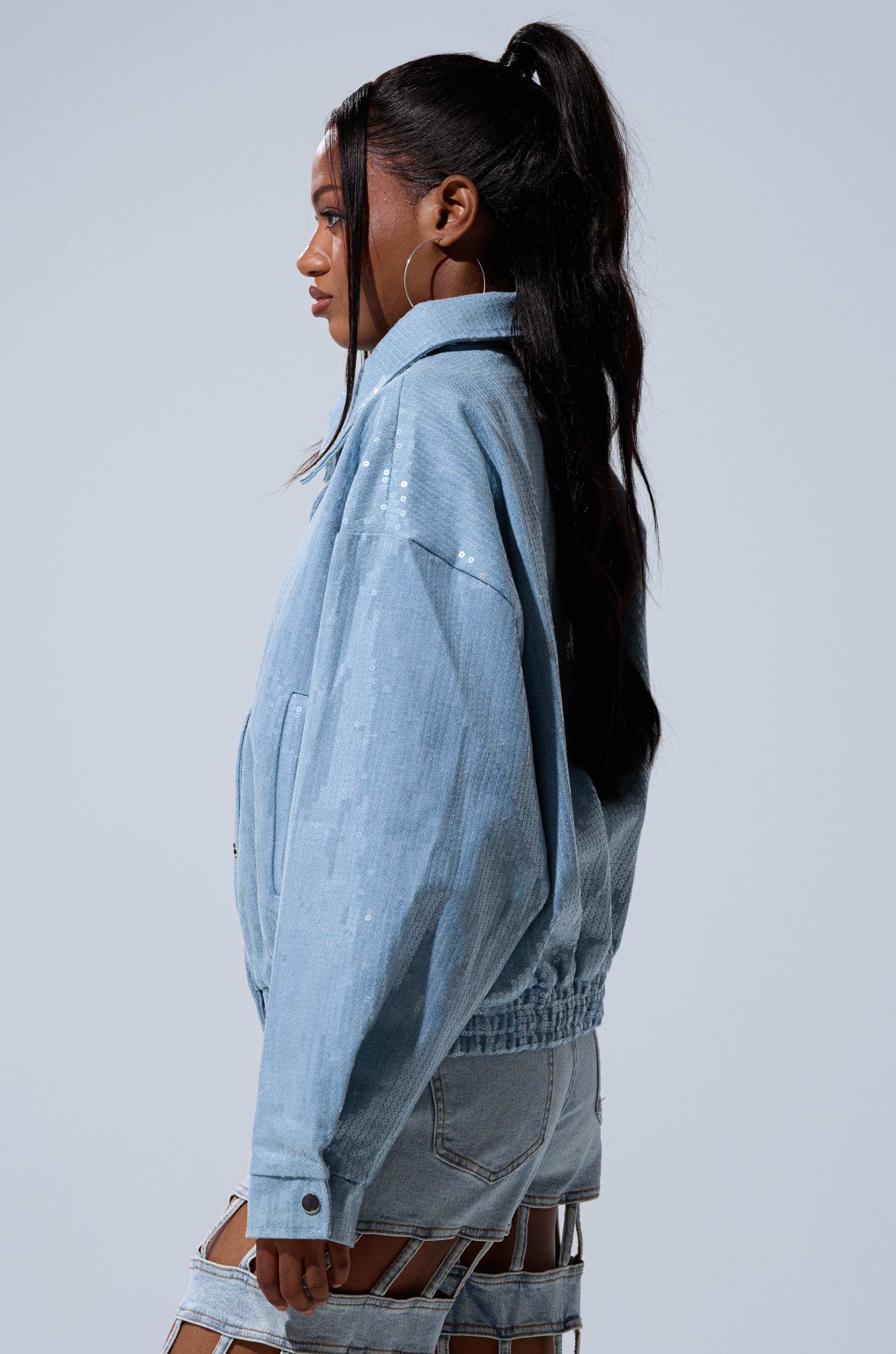 A woman with long, wavy hair in a high ponytail models the SEQUIN OVERLAY DENIM BOMBER and distinctive denim shorts with cut-out details, posing in profile against a plain light background.