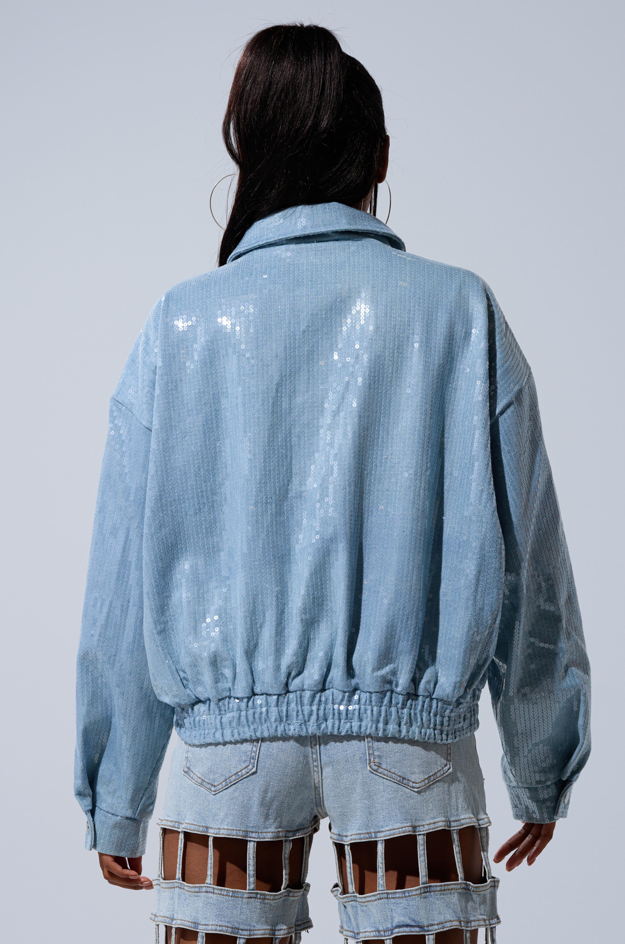 A person with long dark hair is shown from the back in a SEQUIN OVERLAY DENIM BOMBER and distressed shorts with large rectangular cutouts, set against a plain gray background.