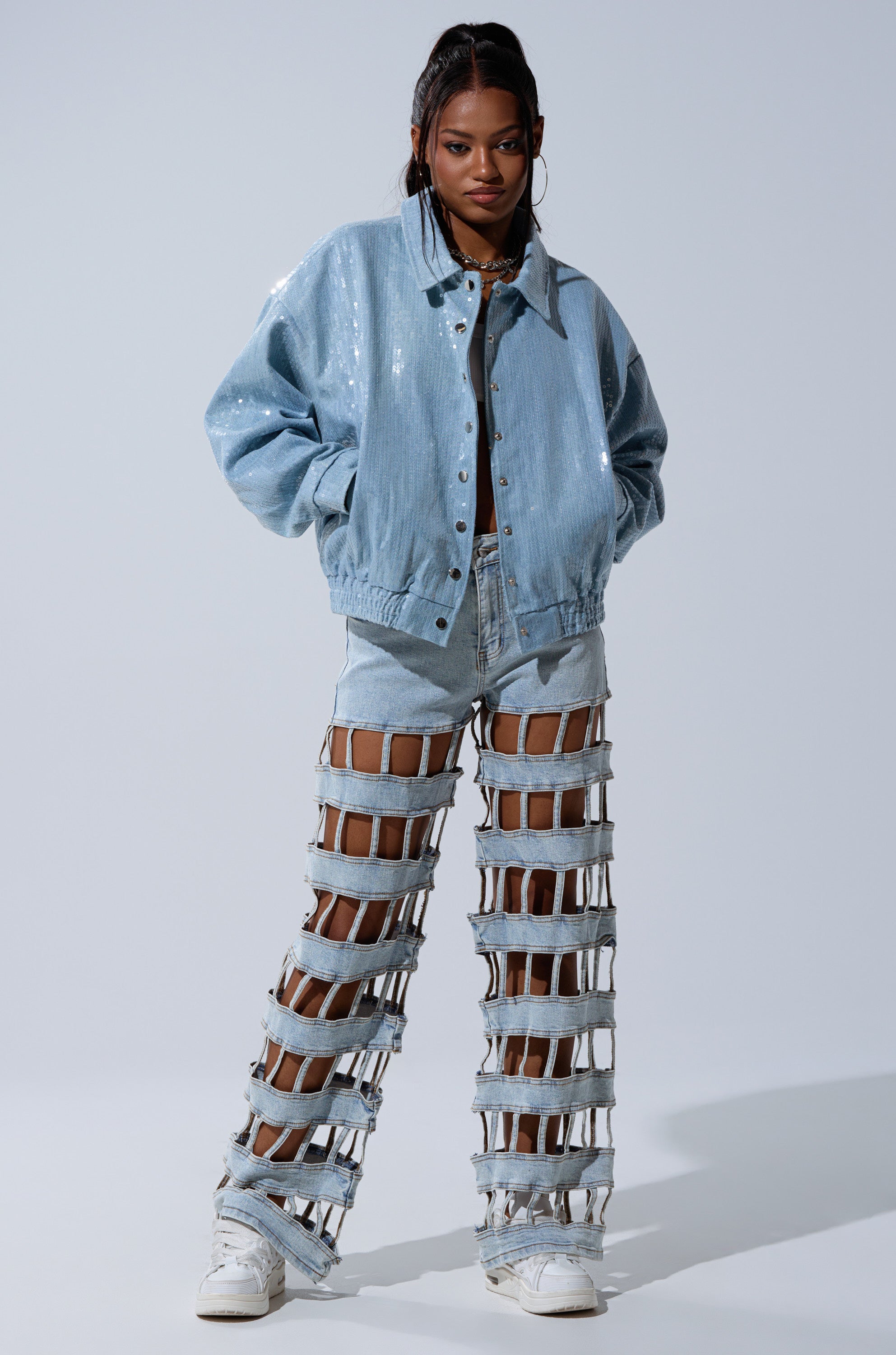 A woman models the SEQUIN OVERLAY DENIM BOMBER, pairing it with bold cutout light blue jeans, white sneakers, and a half-up, half-down hairstyle.