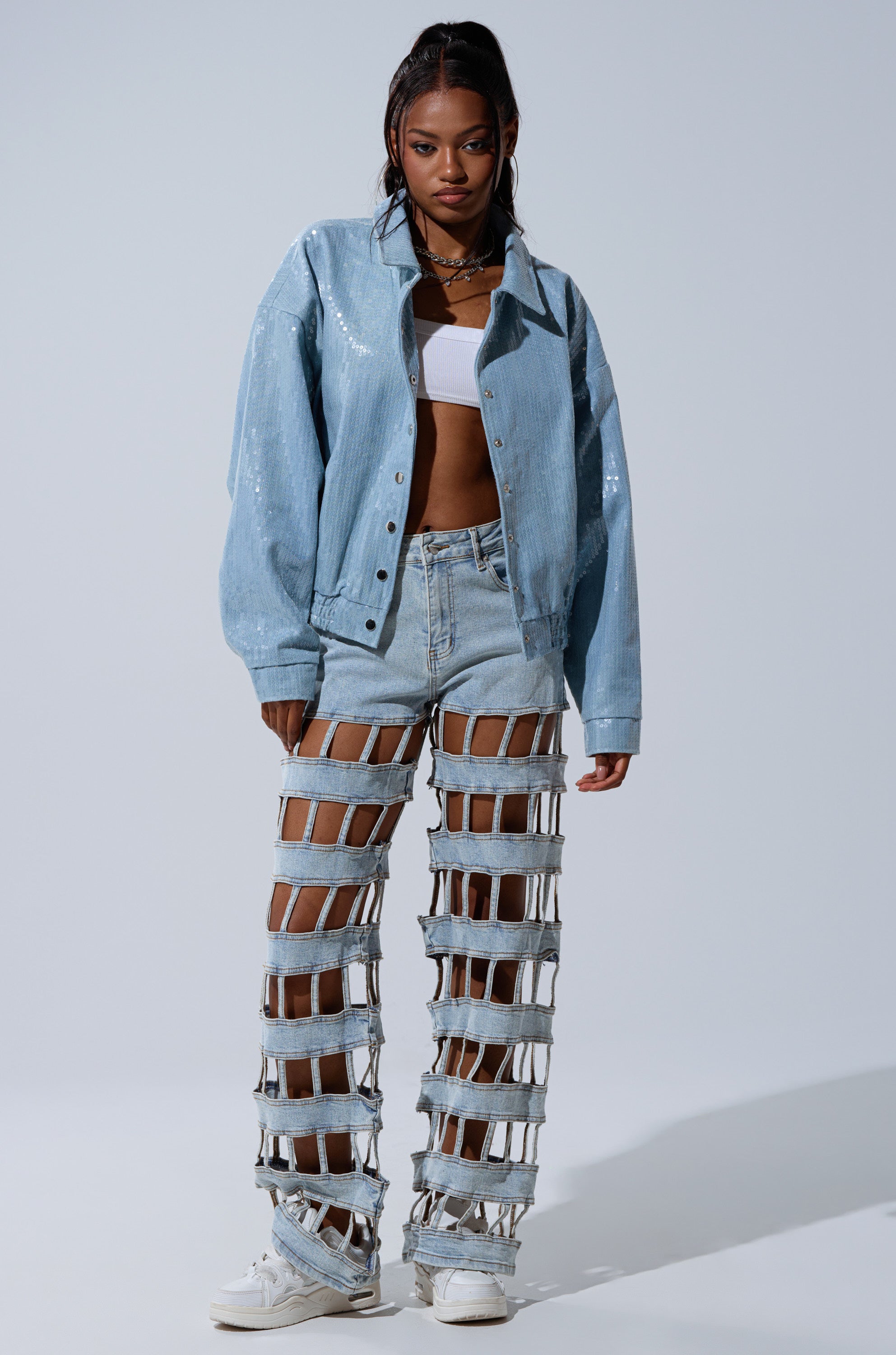 A woman faces forward in a SEQUIN OVERLAY DENIM BOMBER over a white crop top and distinctive cutout denim pants held by straps, with long dark hair and white sneakers, set against a plain backdrop.