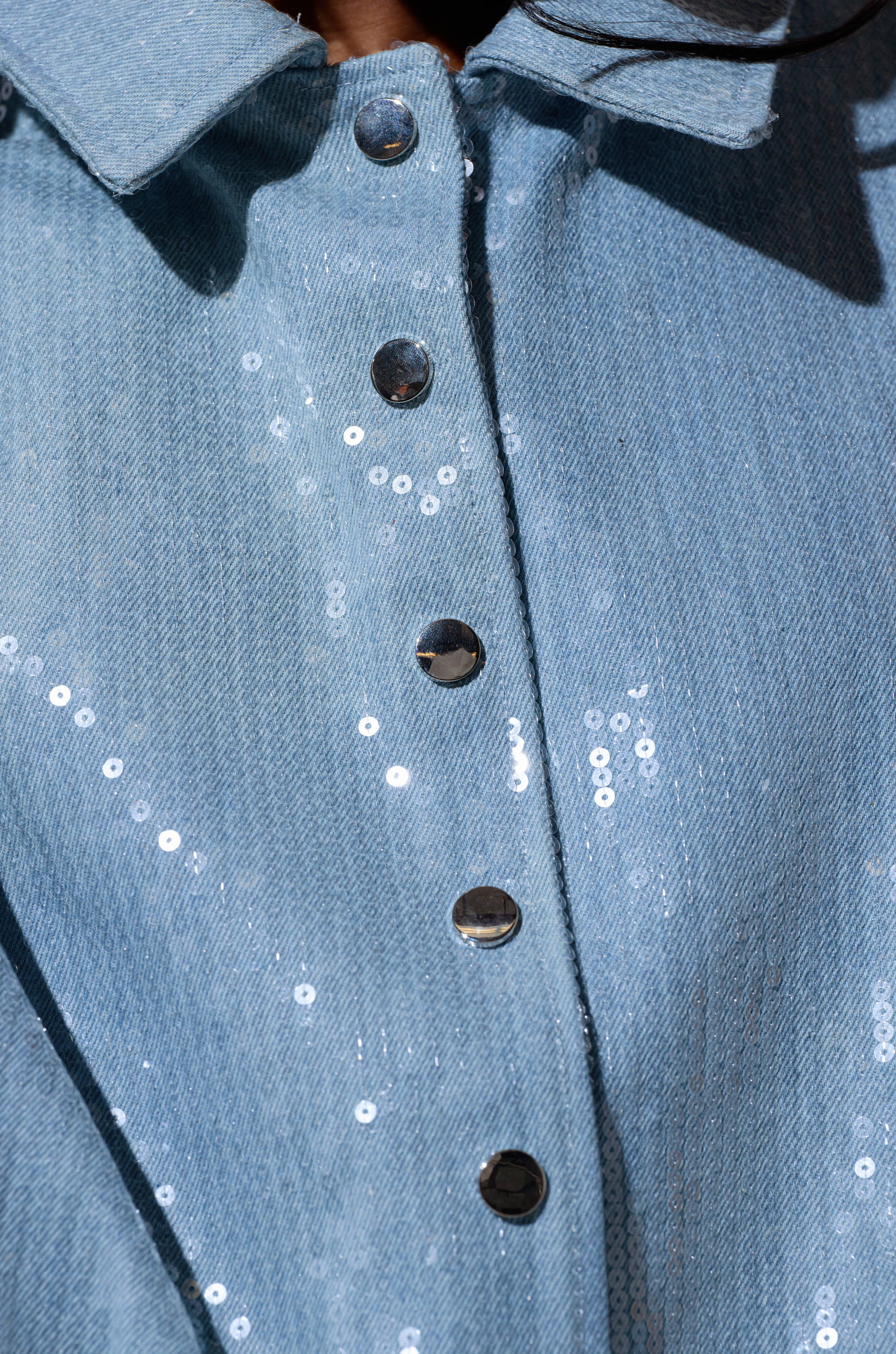 Close-up of the SEQUIN OVERLAY DENIM BOMBER, a light blue denim outerwear shirt featuring shiny sequin accents and metallic front buttons.