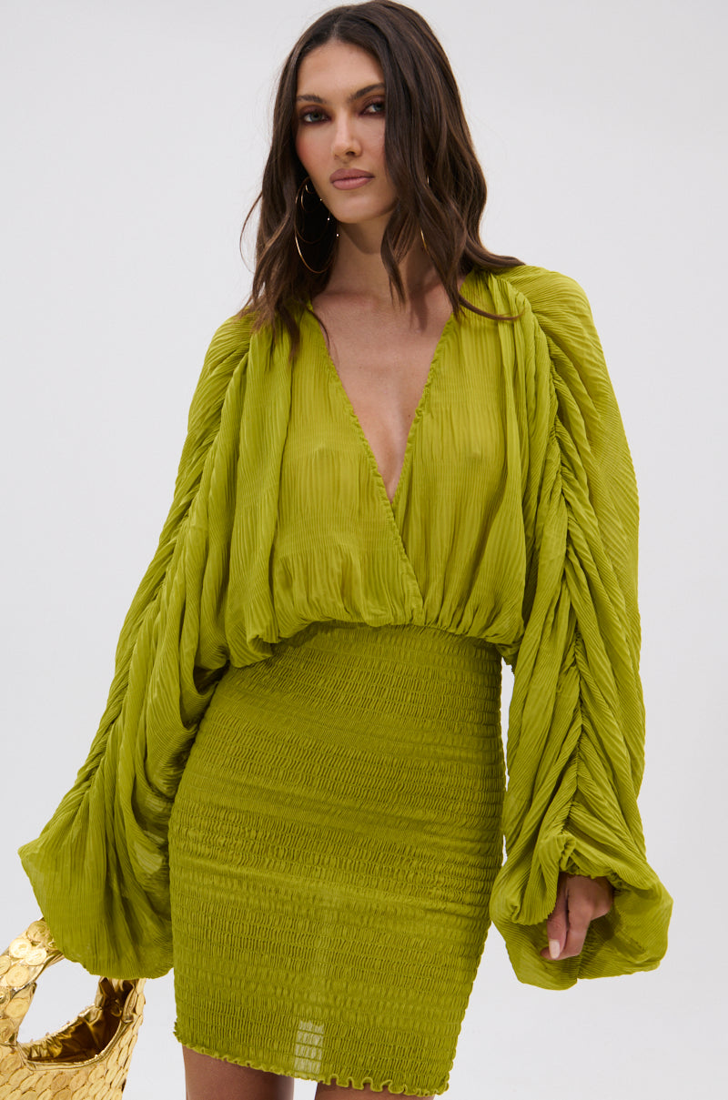 A woman in the NIGHTS LIKE THIS CHIFFON MINI DRESS, featuring a vibrant chartreuse hue, deep V-neck, voluminous long sleeves, and ruched fitted skirt, stands against a plain background holding a gold textured handbag. 1
