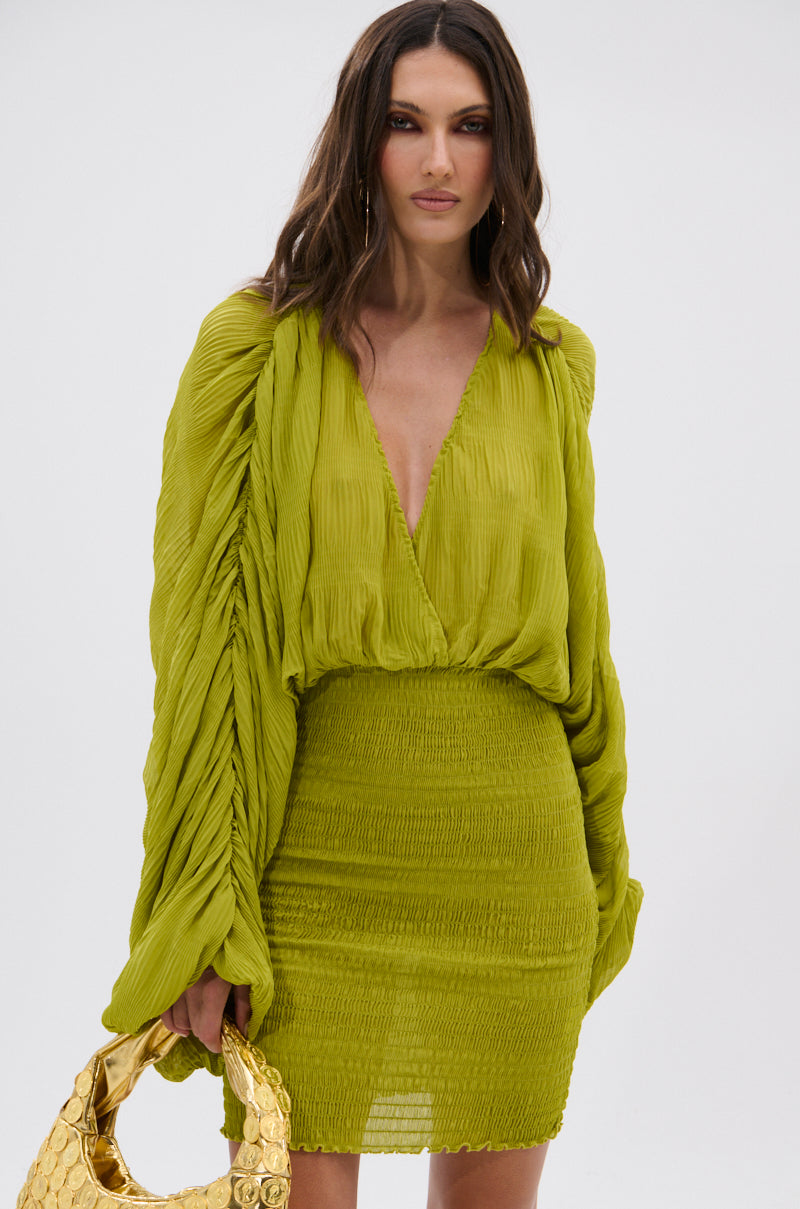 A woman with wavy brown hair wears the NIGHTS LIKE THIS CHIFFON MINI DRESS, featuring long sleeves, a deep V-neck, and a textured skirt. She holds a gold handbag and stands against a plain white background. 2