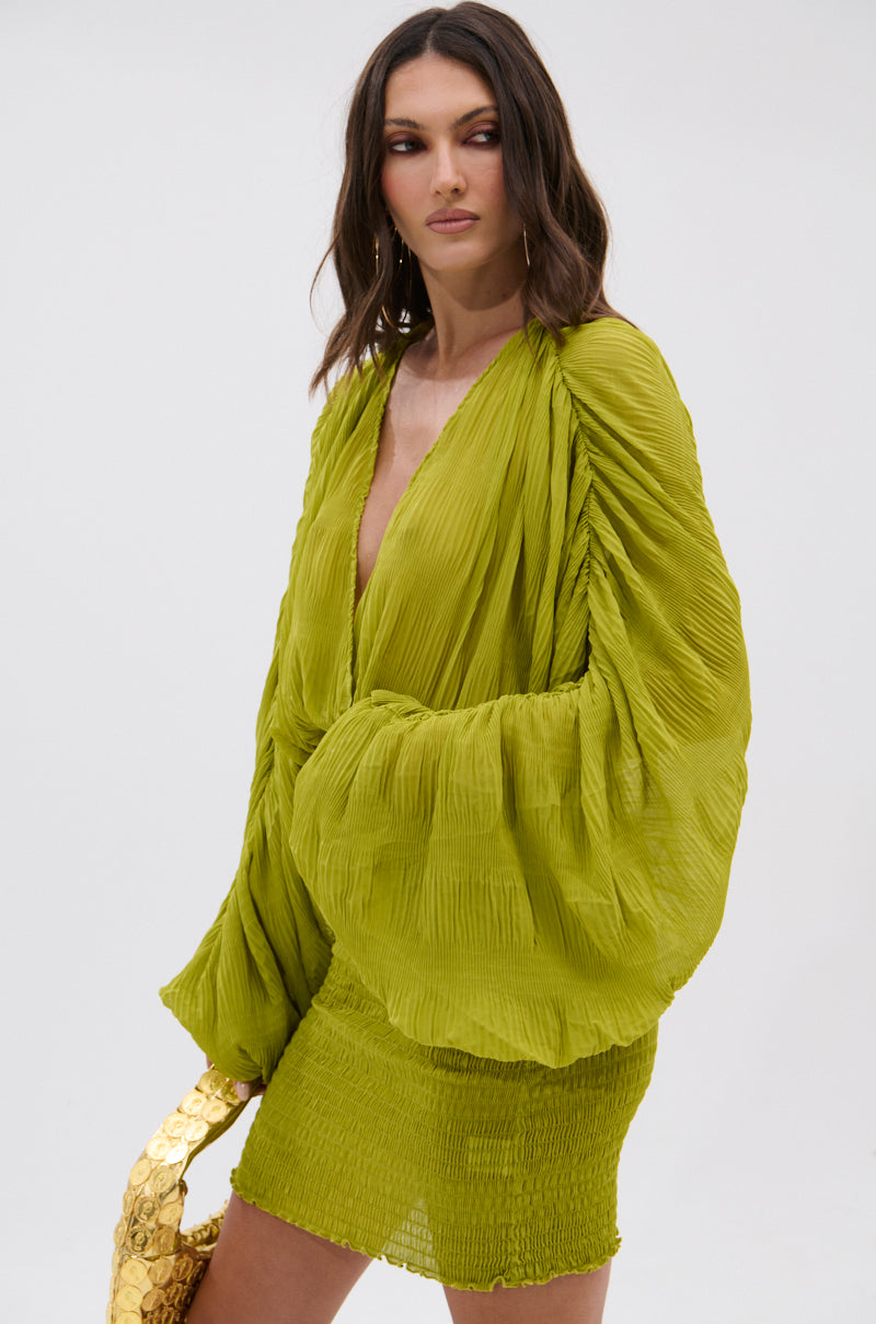 A woman stands against a plain light background, wearing the NIGHTS LIKE THIS CHIFFON MINI DRESS in vibrant green with billowy sleeves and holding a shiny gold bag, evoking chic chartreuse style. 3