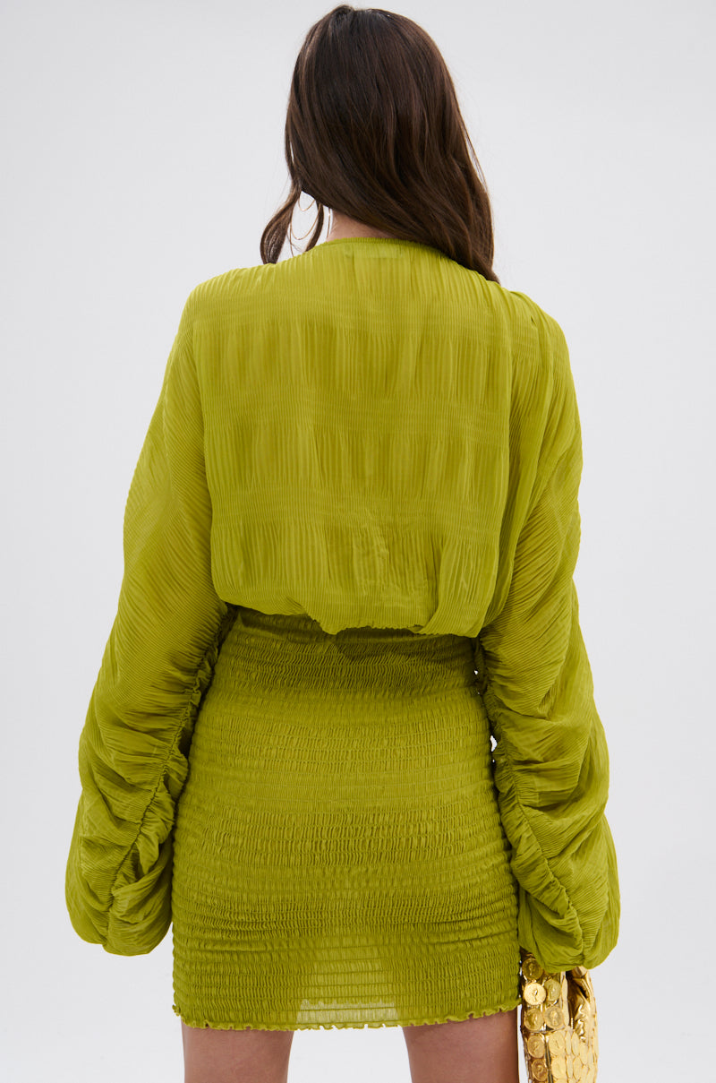 A woman with long brown hair, facing away, wears the NIGHTS LIKE THIS CHIFFON MINI DRESS in chartreuse with ruched long sleeves and holds a gold handbag, set against a plain light background. 4