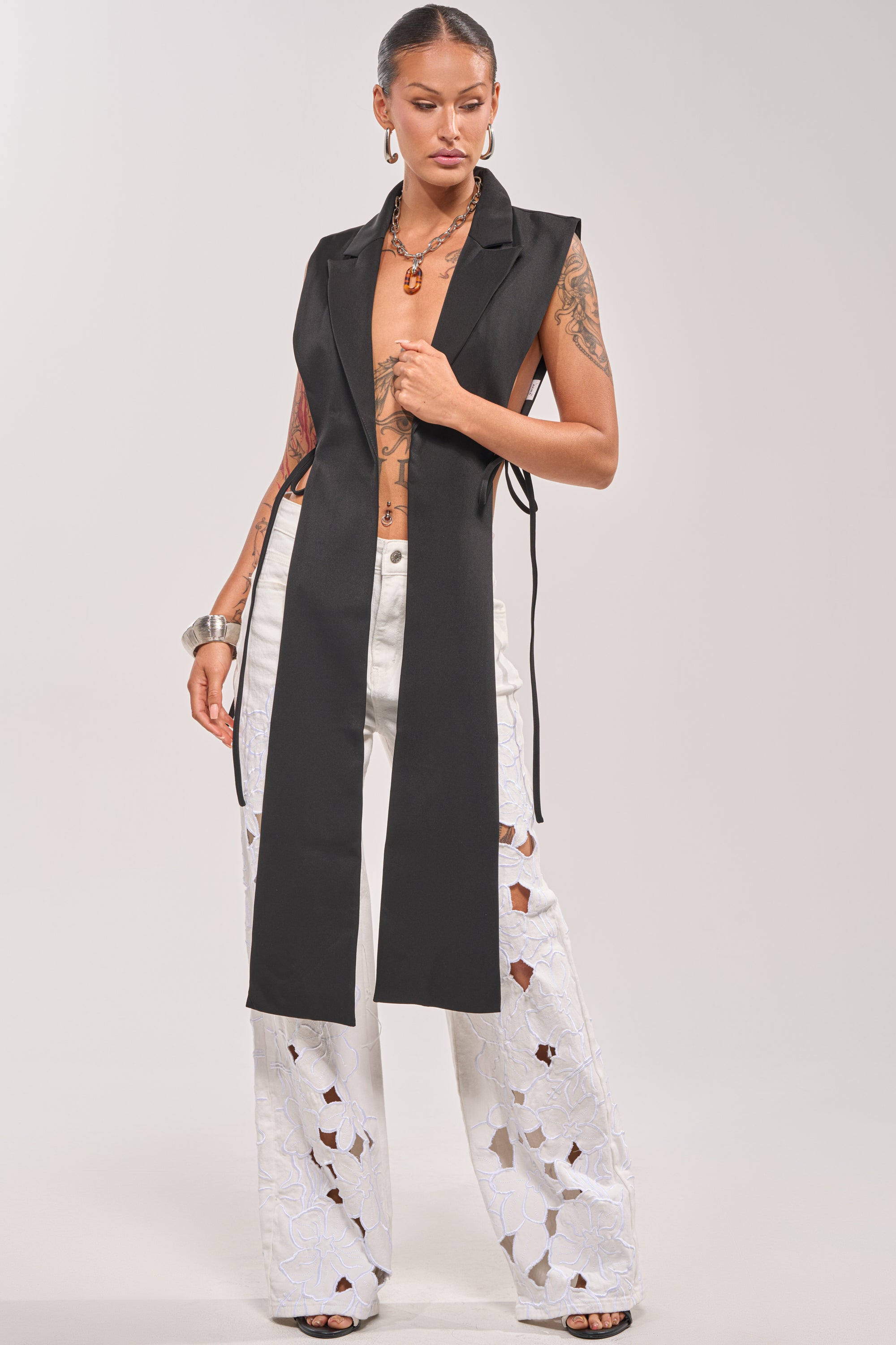 A woman poses confidently in white, wide-leg pants with floral cutouts and the HONEY HONEY TIE VEST IN BLACK, accessorized with chunky jewelry and her hair pulled back to reveal tattoos on her arms. 0