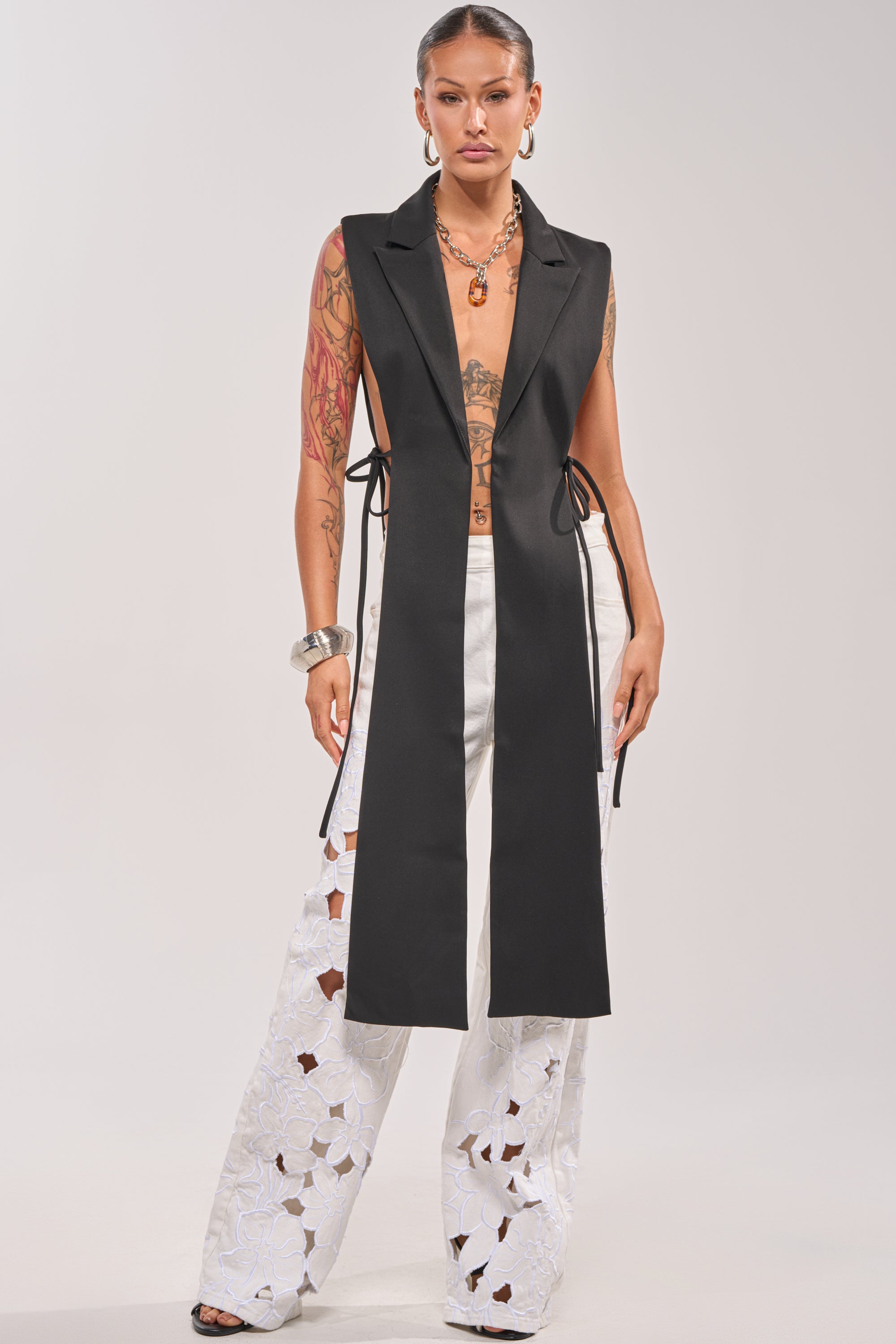 A woman faces forward in the HONEY HONEY TIE VEST IN BLACK, an asymmetrical piece with deep lapels and a high-low hem, styled over white wide-leg floral cutout pants. She accessorizes with hoop earrings, a necklace, tattoos, and slicked-back hair. 1