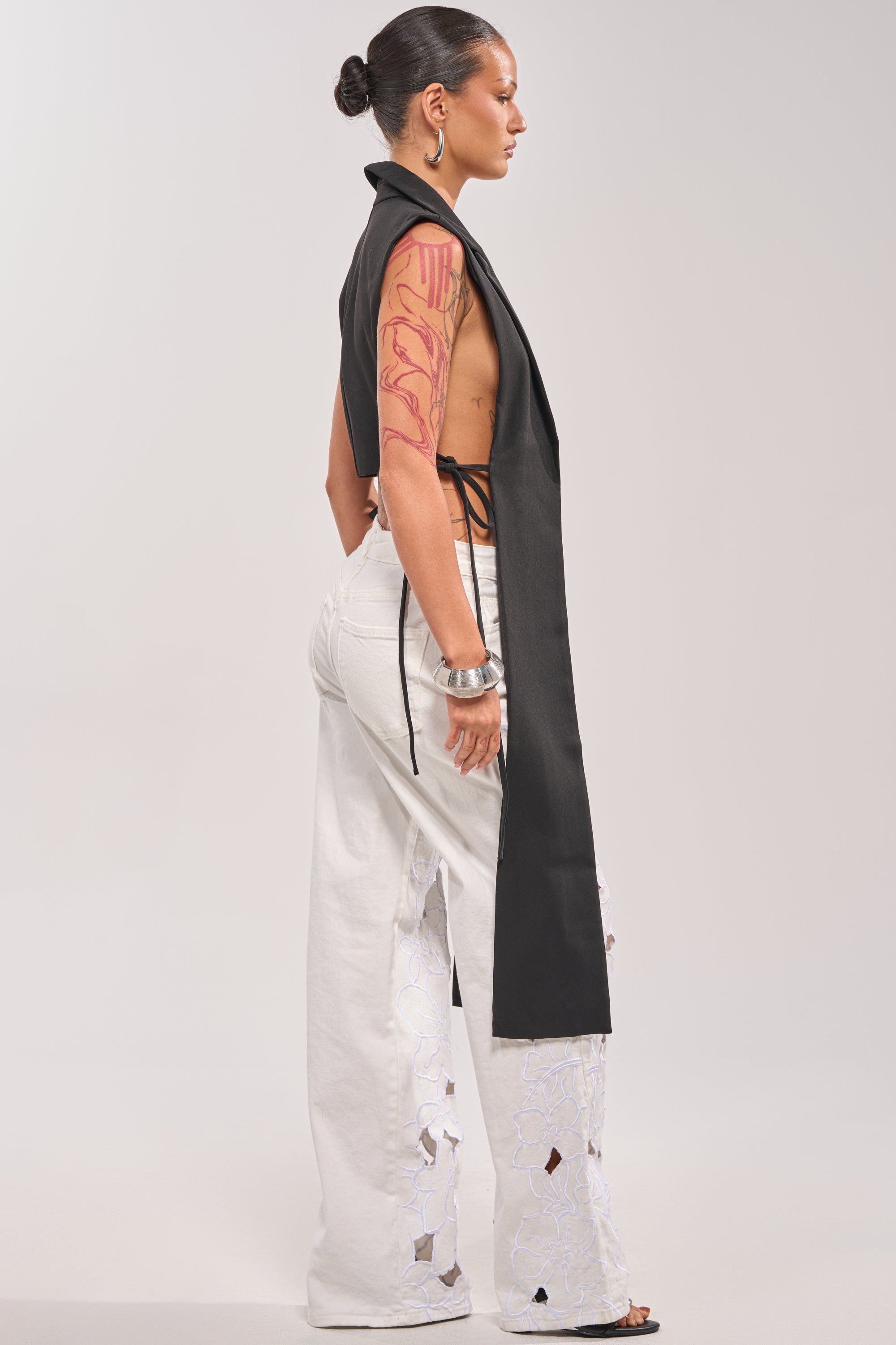 A person with a tattooed arm and slicked-back hair models the HONEY HONEY TIE VEST IN BLACK over a bare back, styled with white wide-leg pants featuring cut-out details, standing sideways against a plain background. 2