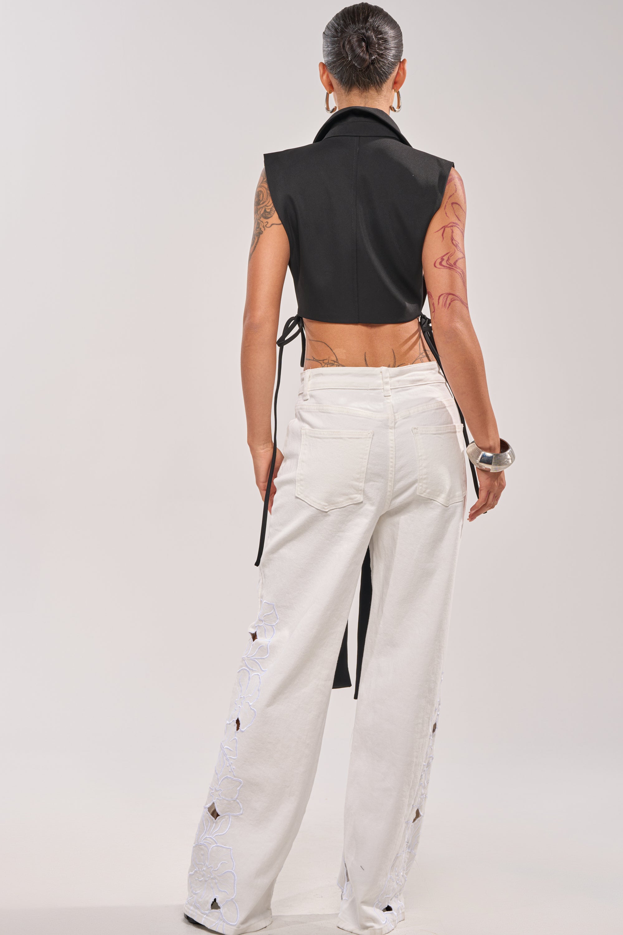 A person with a bun faces away, wearing the HONEY HONEY TIE VEST IN BLACK, white wide-leg jeans with holes and floral patterns, silver earrings, a chunky bracelet, and showing tattoos on their left arm. 3