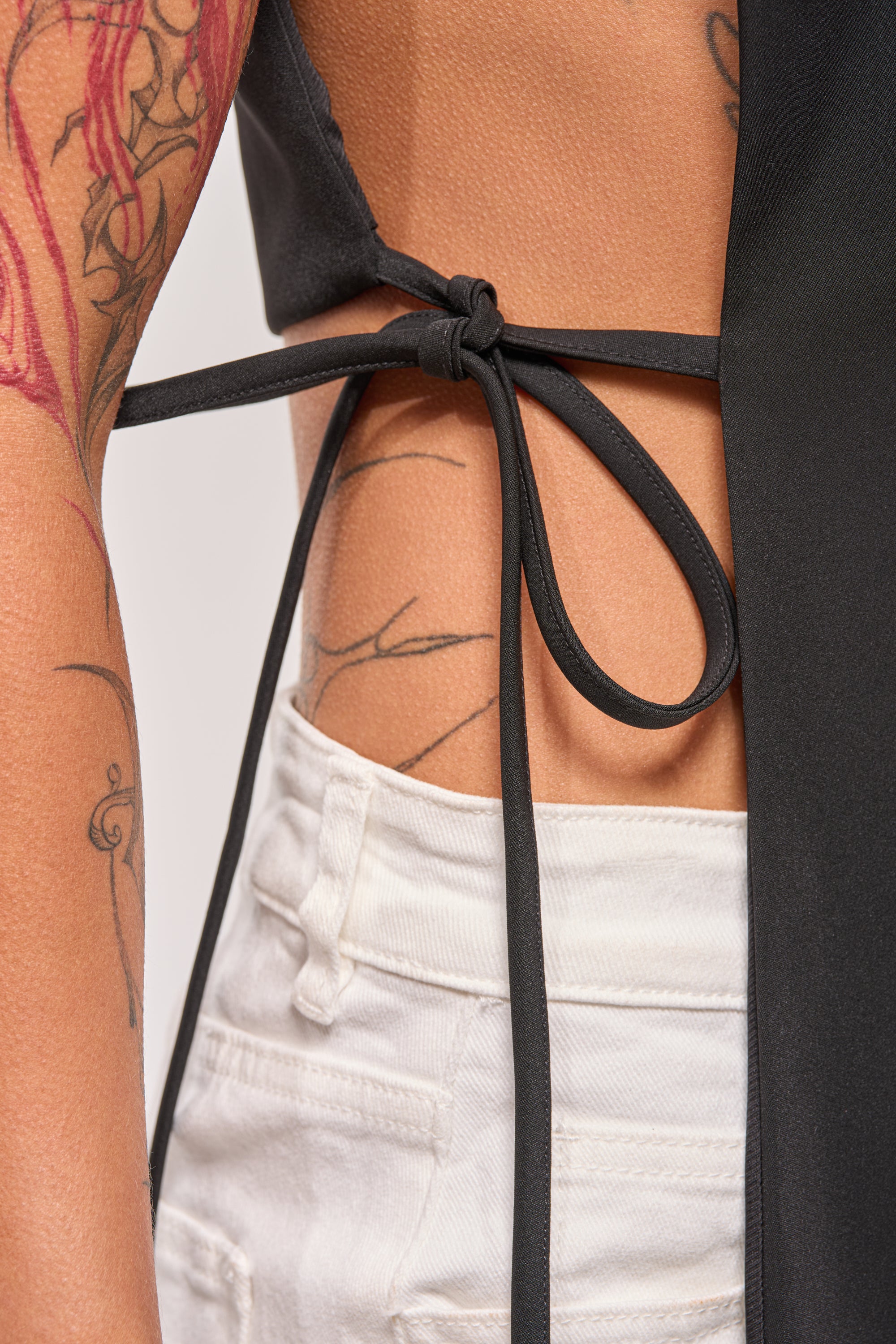 Close-up of a person's side wearing the HONEY HONEY TIE VEST IN BLACK, tied at the back with strings over white high-waisted pants, showcasing tattoos on their arm and torso. 4