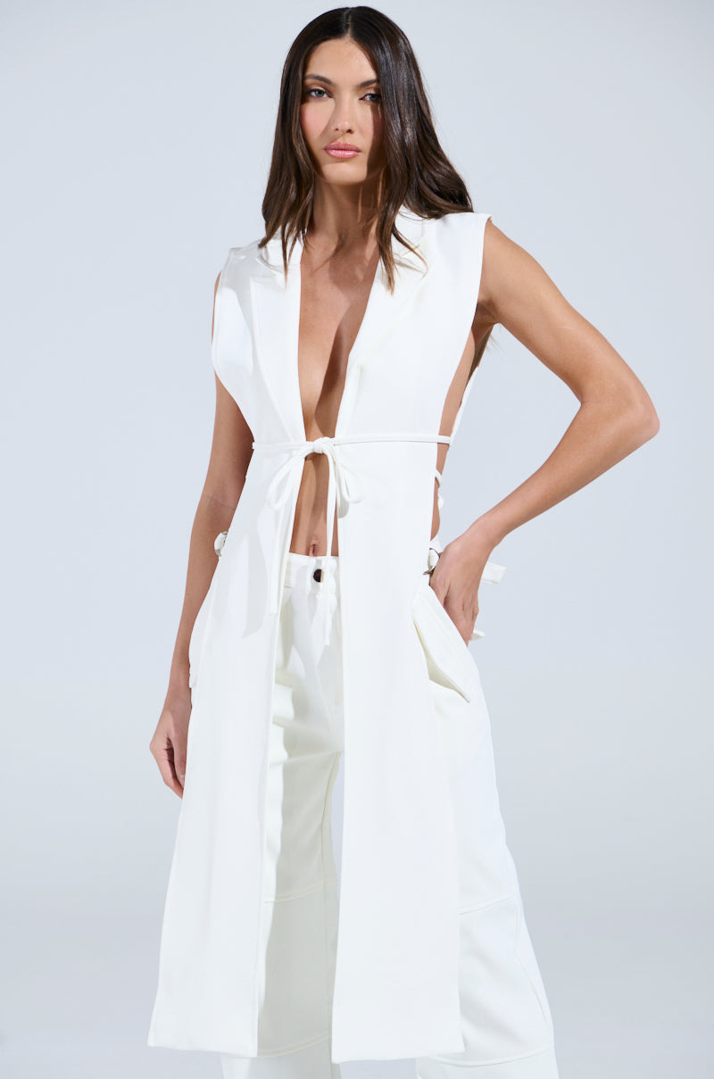 Wearing the chic HONEY HONEY TIE VEST by AKIRA Label, paired with matching high-low silhouette pants, their long, straight hair flows gracefully against a plain light background.