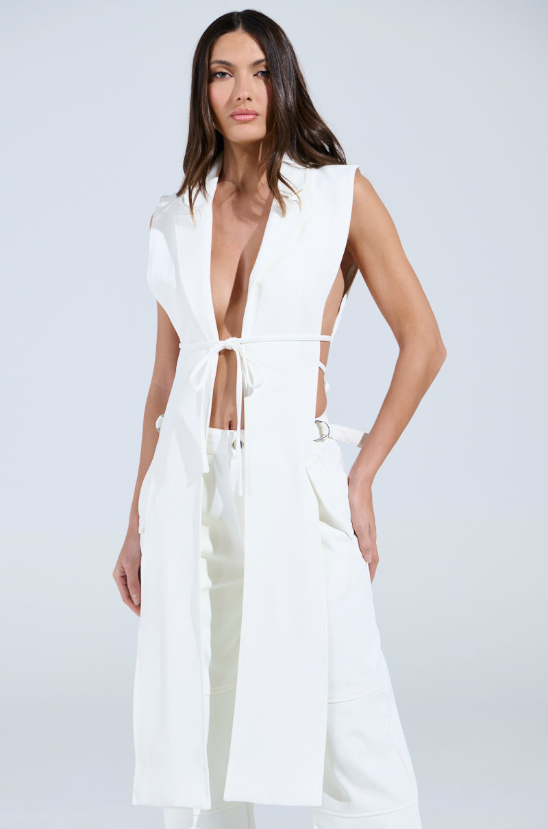 Wearing a stylish sleeveless HONEY HONEY TIE VEST with a deep V-neck and tie-up front, paired with matching wide-legged pants, the person stands against a plain backdrop. With one hand in the pocket and long, dark hair flowing, they exude confidence.